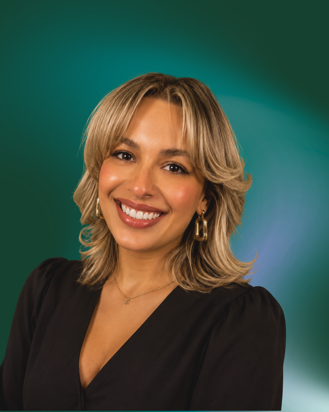 A woman with shoulder-length wavy blonde hair, smiling with white teeth, wearing a black top with puffed sleeves, gold hoop earrings, and a delicate necklace, against a green and blue blurred background.