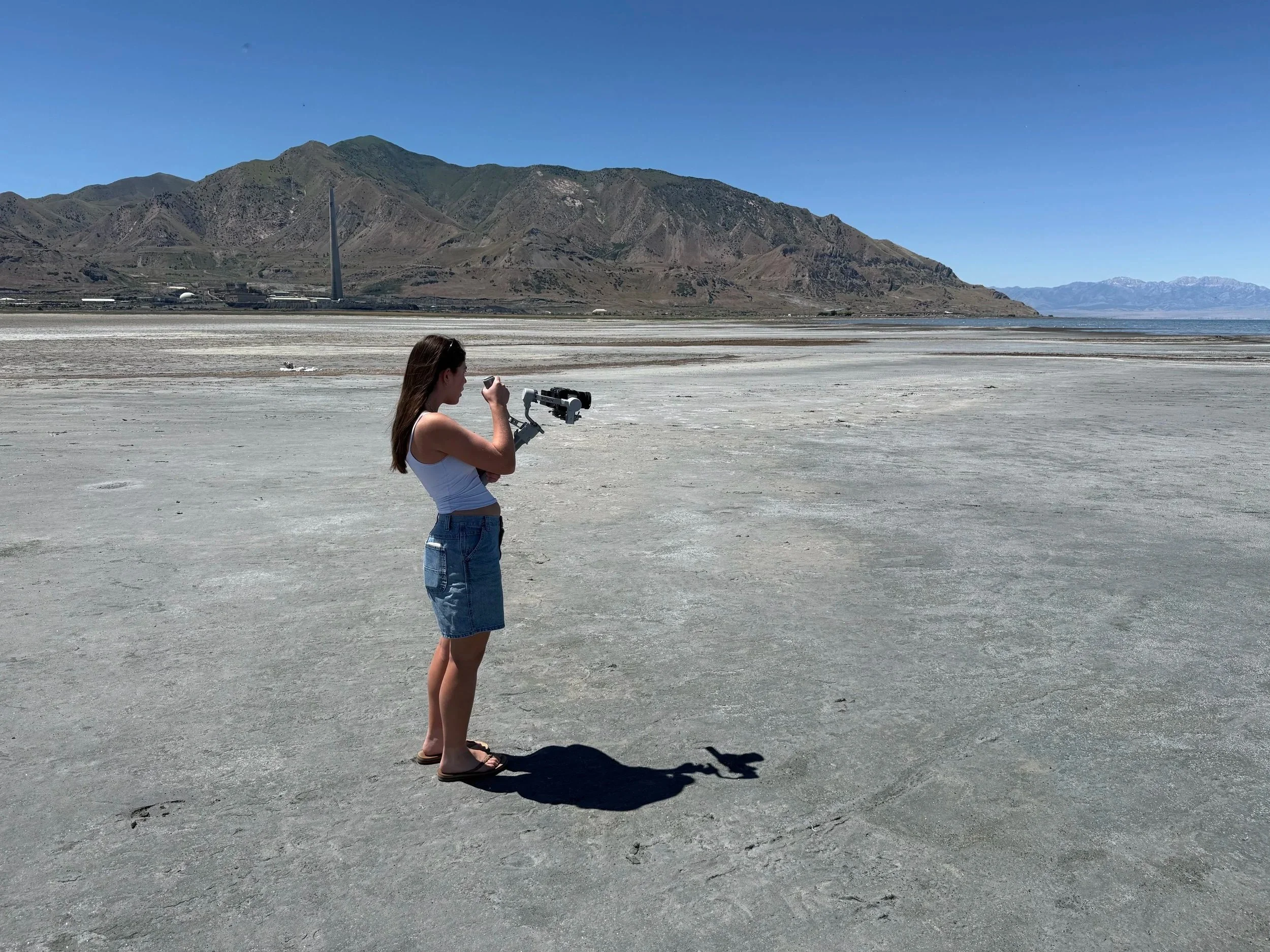 A woman in a white tank top and denim shorts is standing on a flat, dry, white landscape, looking through a telescope. She is barefoot, and the landscape extends to distant mountains under a clear blue sky.