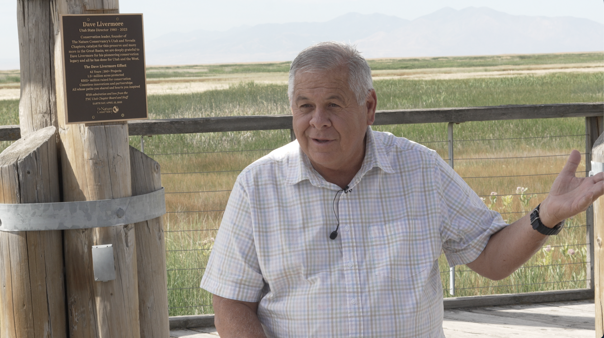 A man with gray hair, wearing a short-sleeved checkered shirt, gestures with his right hand while speaking outdoors. Behind him is a wooden fence, a metal gate, and a landscape of green fields and distant mountains. There is a plaque on the wooden post, honoring Dave Livermore, Utah State Director from 1980 to 2023, about conservation efforts.