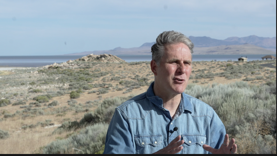 man in denim shirt speaking outdoors near a body of water with hills and open land in the background