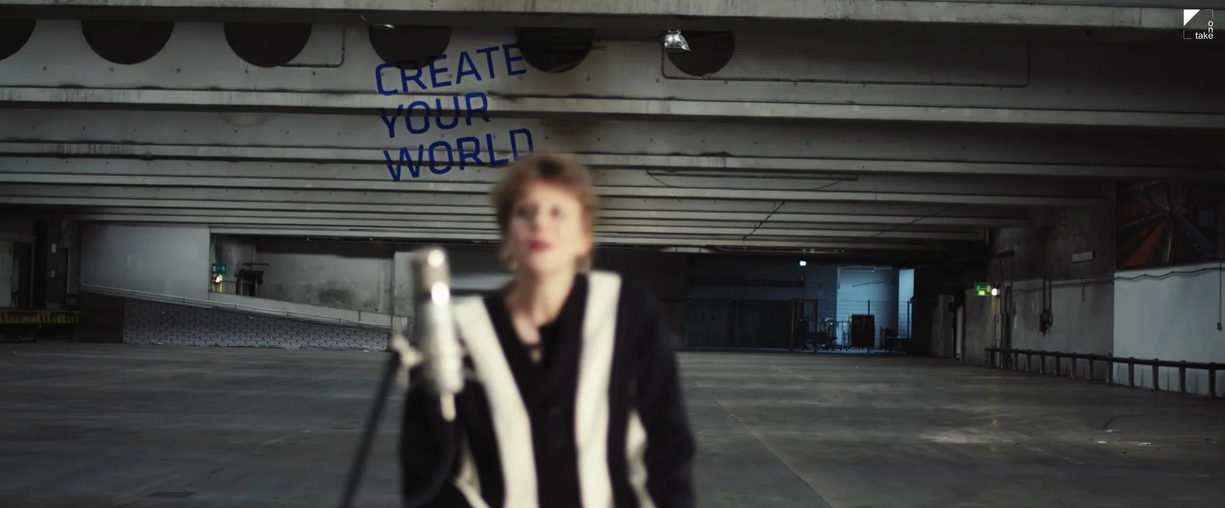 Unscharfe Person in schwarzer Kleidung mit weißen Streifen in einer verlassenen Industriehalle; dahinter steht „CREATE YOUR WORLD“.
