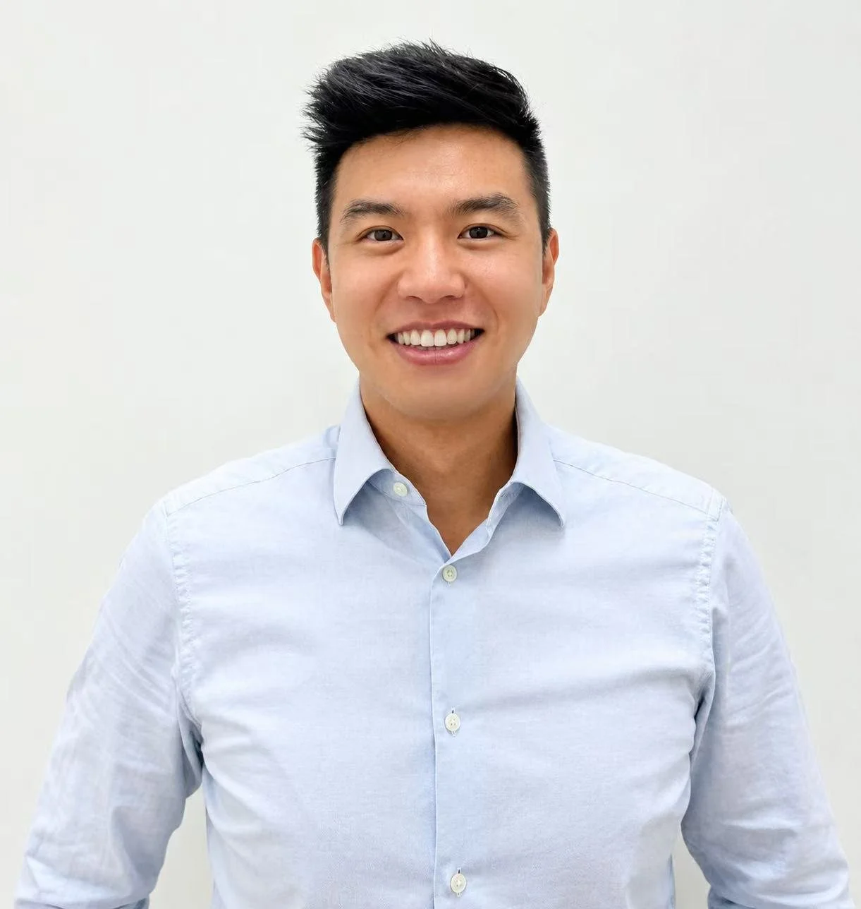 Smiling young Asian man in a light blue button-up shirt standing against a plain white background.