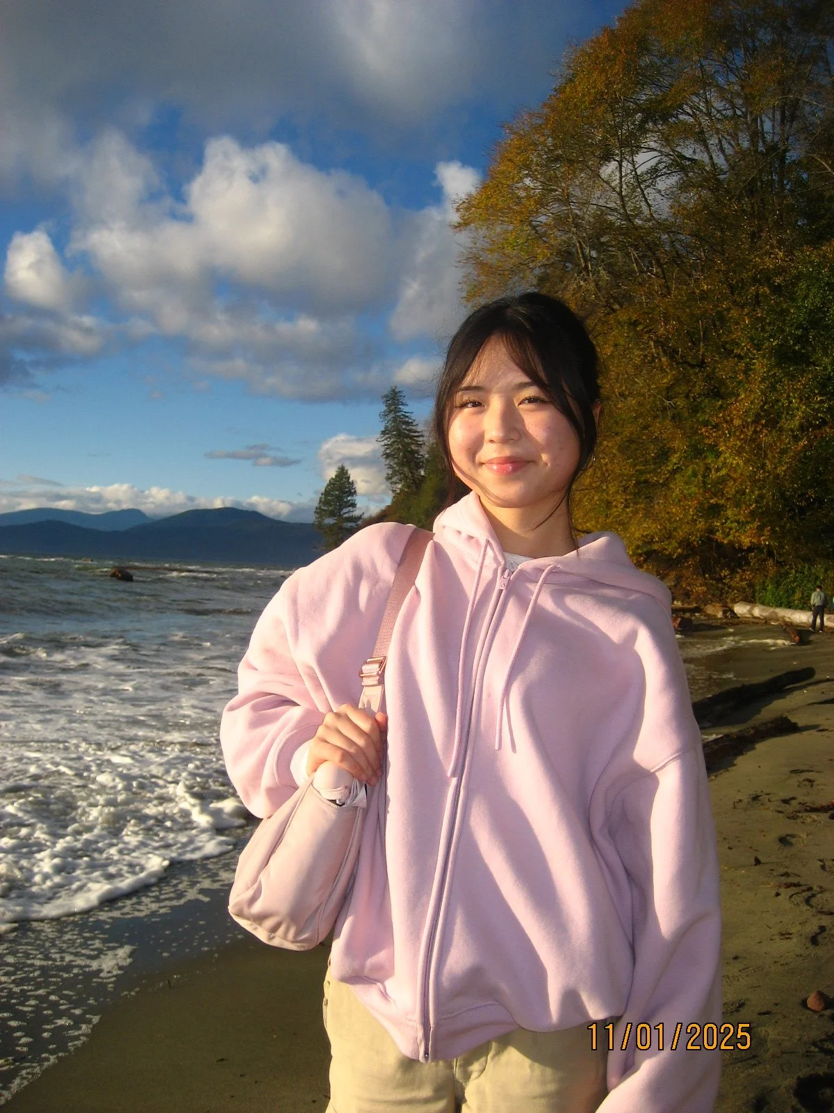 A young woman standing on a beach with ocean waves, mountains in the background, and trees with autumn foliage, wearing a pink hoodie and smiling.