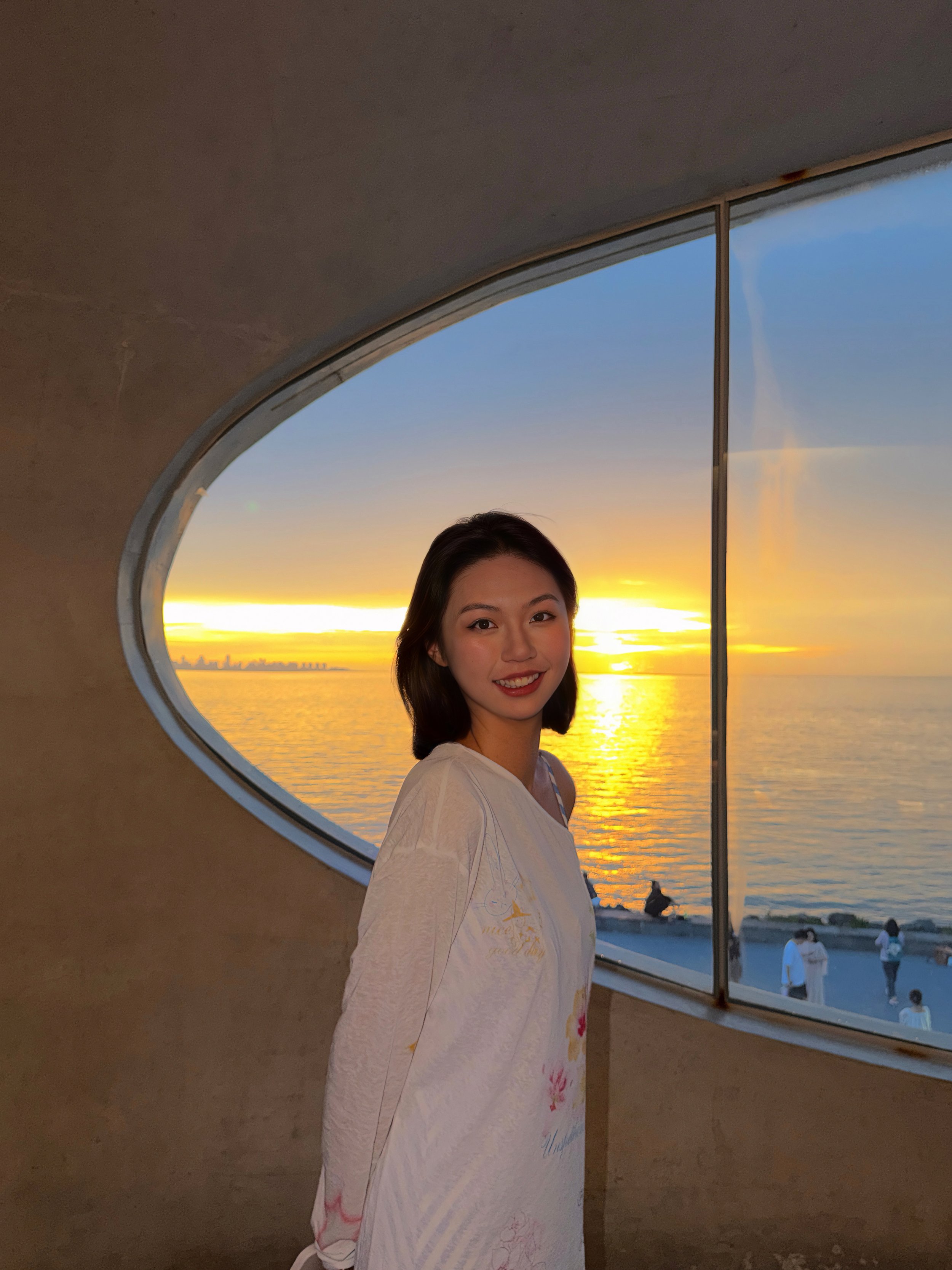 A woman smiling inside a building with a large circular window showing a sunset over water.