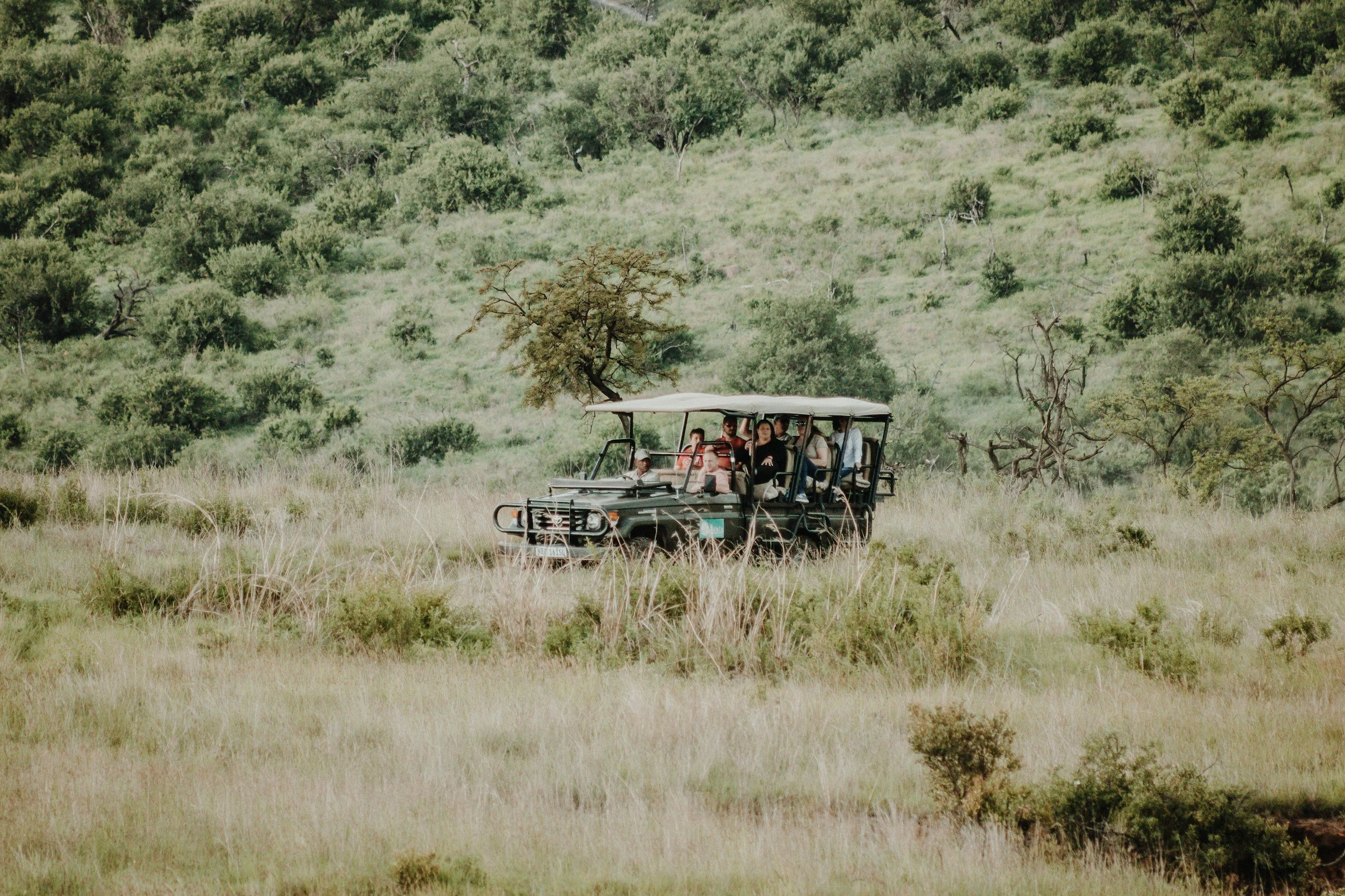 A group of people riding an open-sided safari vehicle through a grassy savanna with scattered trees.