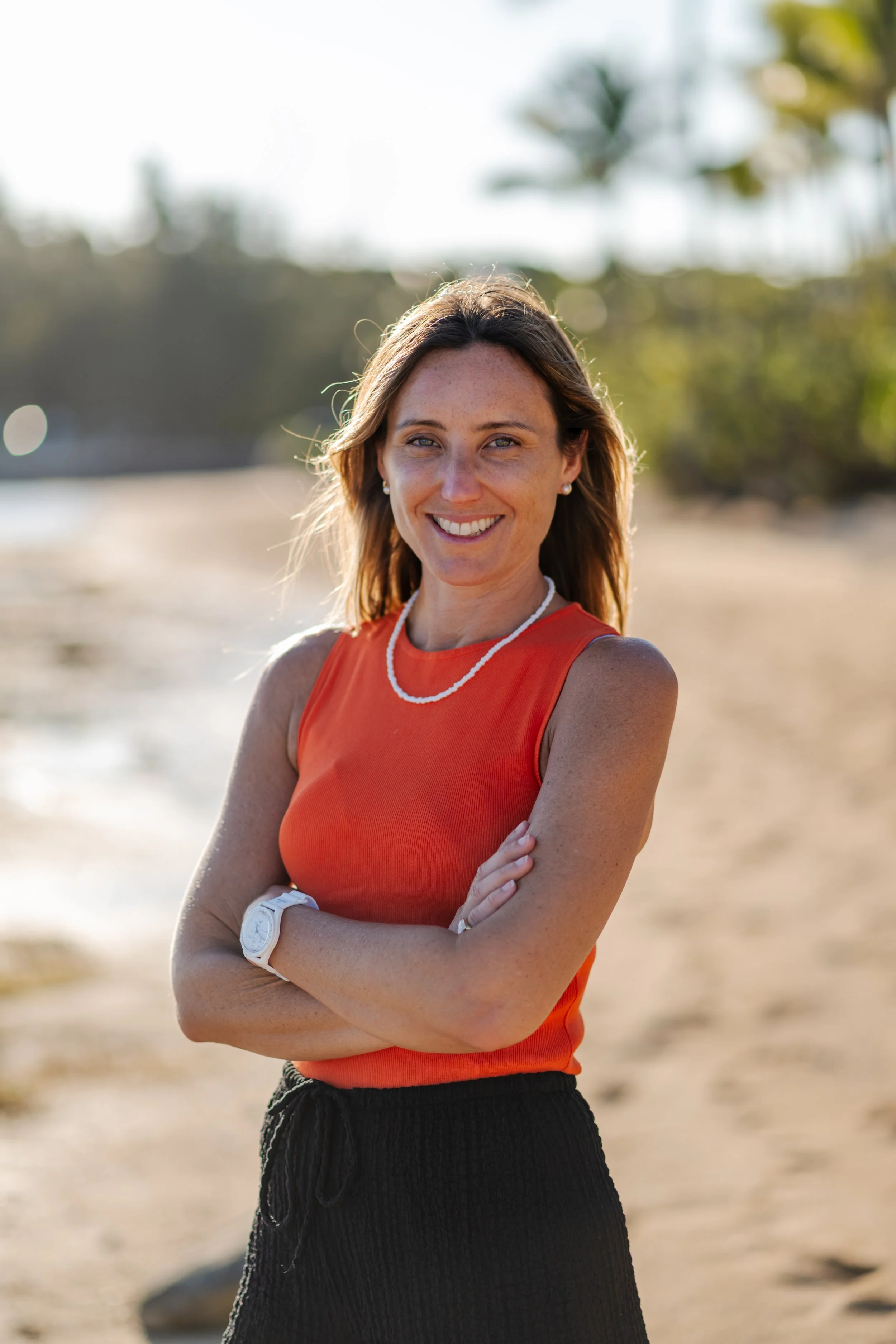 A woman with light skin and brown hair standing on a beach, smiling, wearing an orange sleeveless top, black pants, a white wristwatch, pearl earrings, and a white necklace, with tropical trees and blurred background.