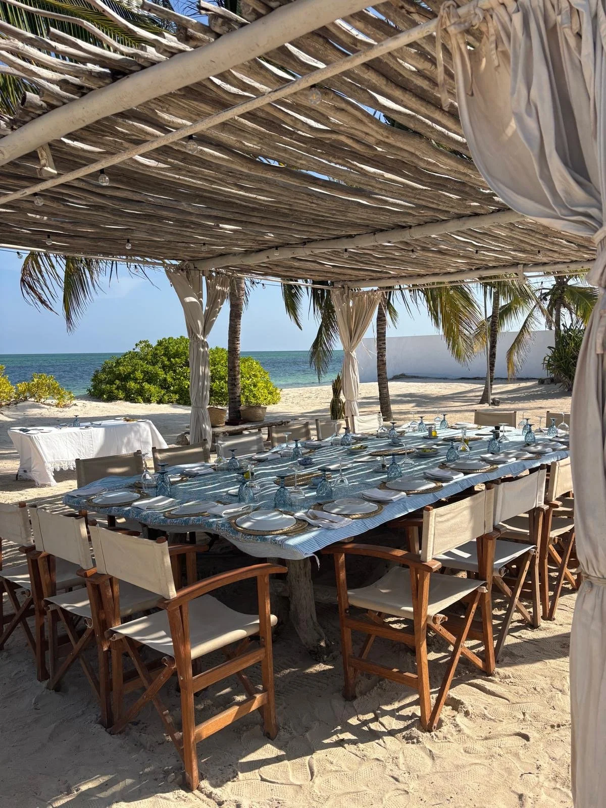 A beachside dining setup with a long table set for a meal, covered by a wooden and fabric canopy, with palm trees, sand, and the ocean in the background.