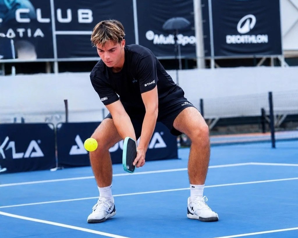 A young man playing pickleball on an outdoor court, ready to hit a yellow ball with his paddle.