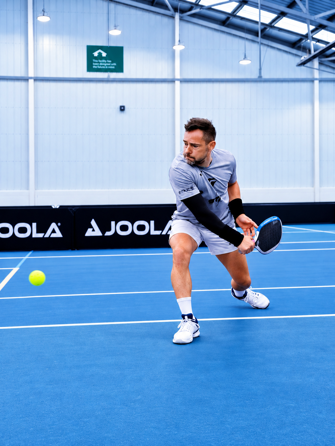 A man playing pickleball indoors in a sports facility with a blue court, holding a paddle, with a pickleball nearby.