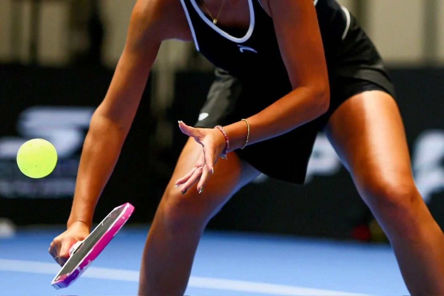 A woman in athletic attire playing pickleball, preparing to hit a yellow ball with a pink and black paddle on a blue court.