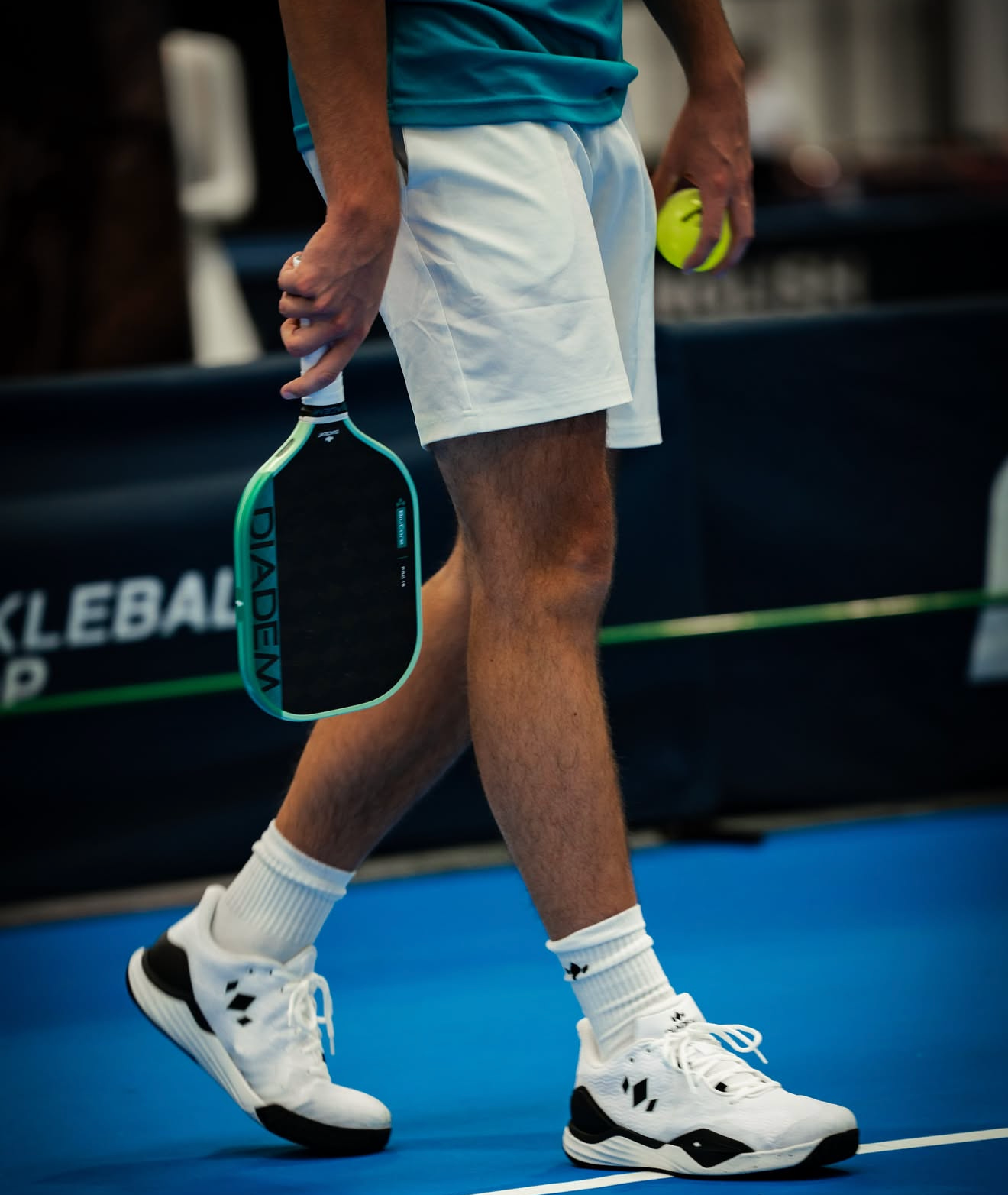 A person wearing white Diadem sports shorts, white socks, and white Diadem pickleball trainers with black accents, holding a yellow pickleball in their left hand and a Diadem paddle in their right hand, on a blue indoor court.