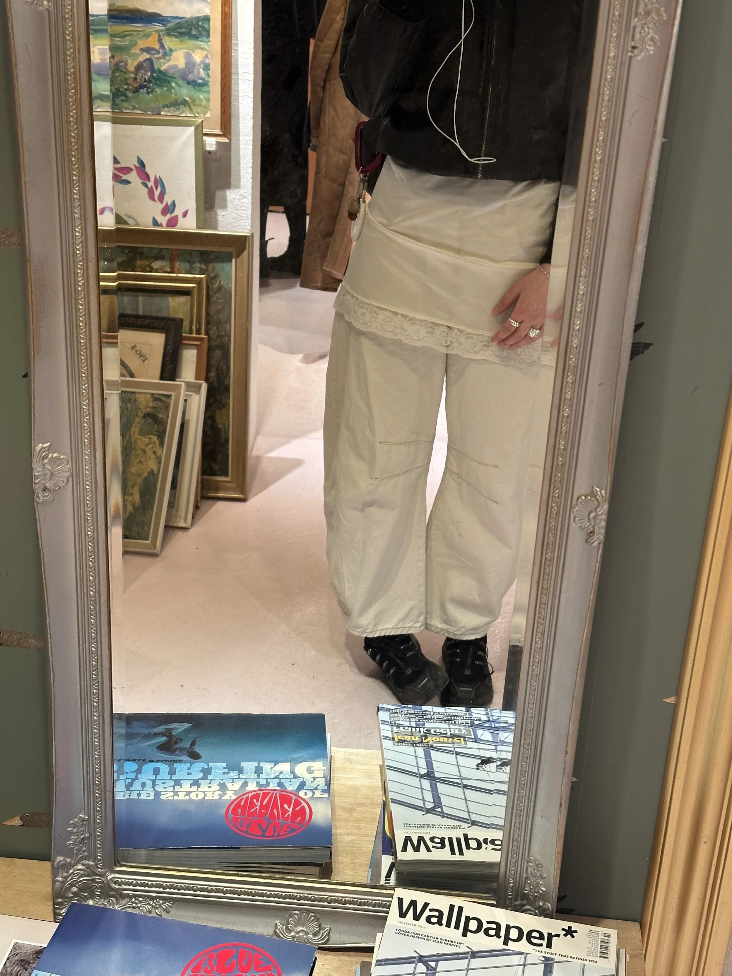 A person taking a mirror selfie in an art store or gallery, wearing black jacket, cream-colored shirt with lace detail, cream pants, and black shoes. The mirror reflects framed artwork on display and various art supplies on a table, with a sign that reads 'Wallpapers' and other items.