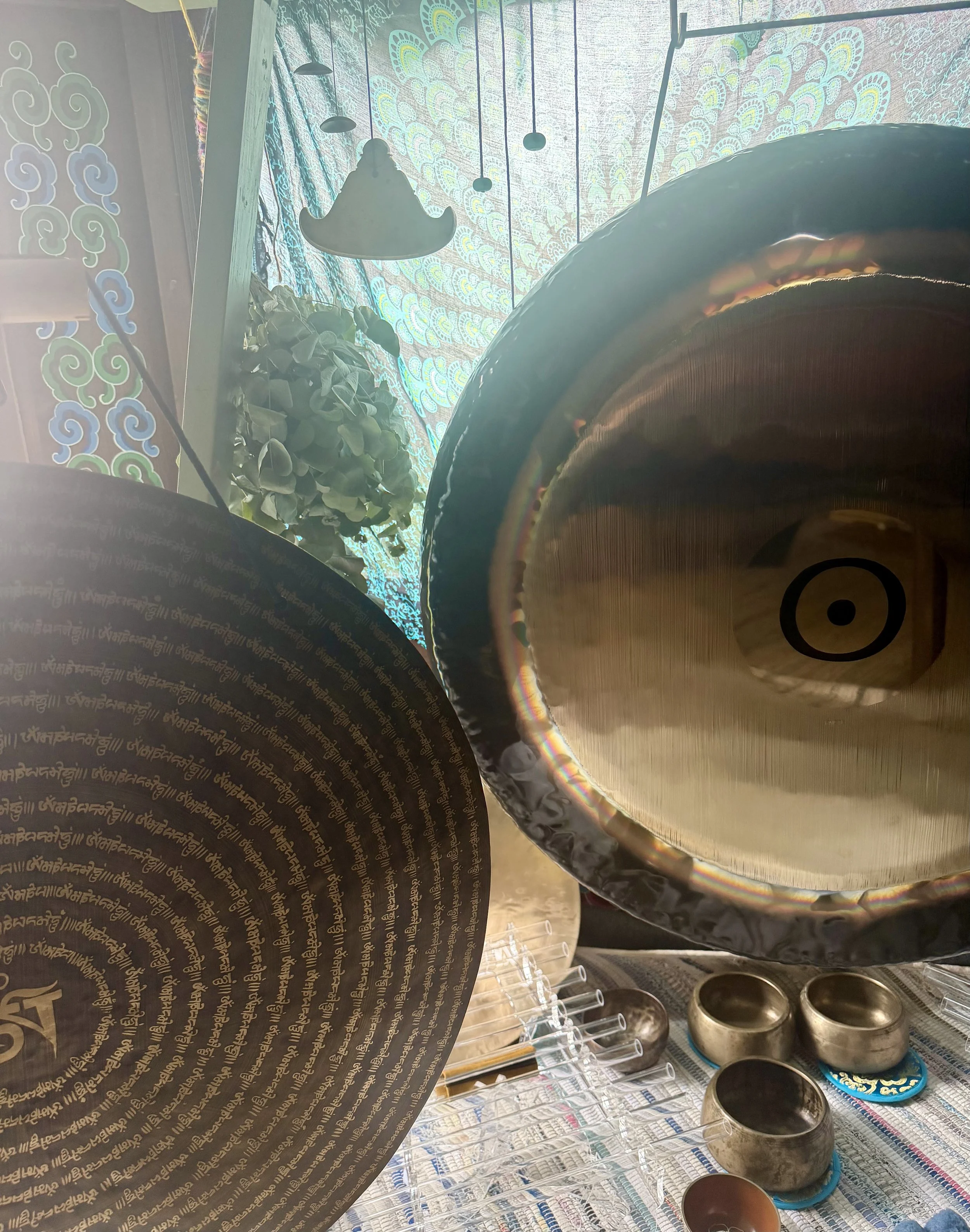 A display of singing bowls of various sizes arranged on a patterned cloth, with a colorful, patterned ceiling, hanging lights, a plant, and a background of a decorated wall with blue, green, and purple designs.