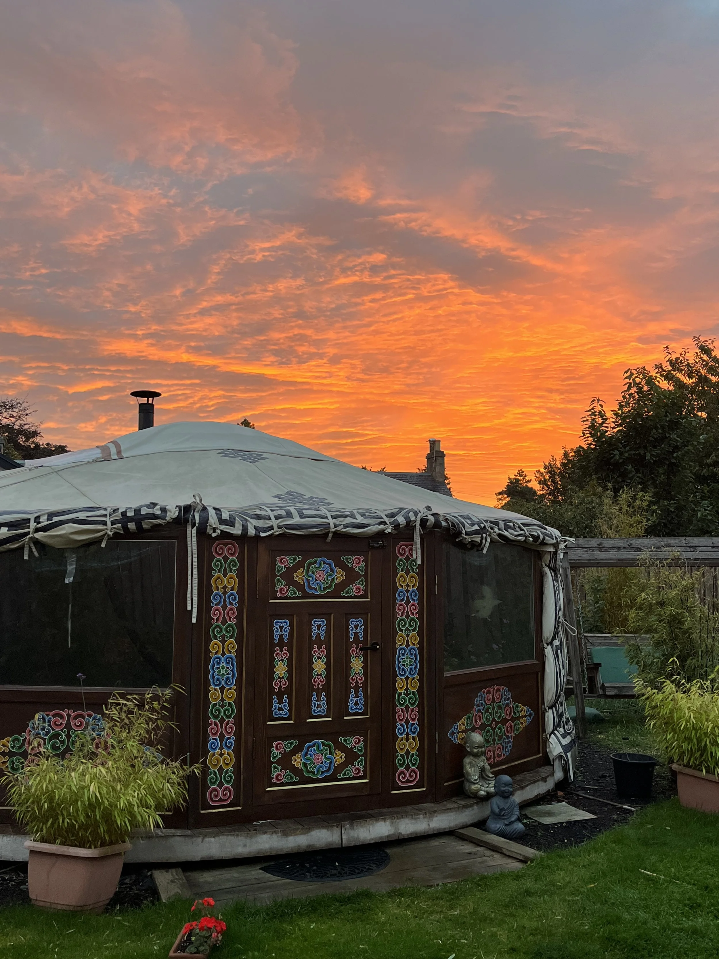 A small wooden structure with colorful decorative patterns, set in a garden during sunset. The sky has orange and pink clouds, and there are potted plants, a Buddha statue, and garden furniture nearby.