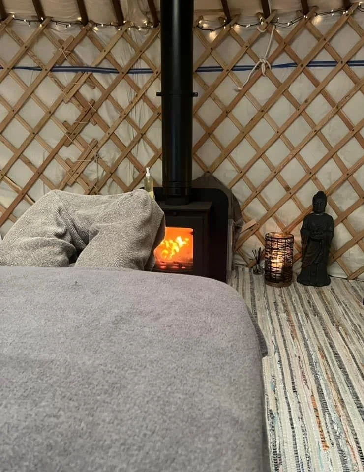Cozy interior scene with a wood stove emitting a warm glow, a gray sofa in the foreground, decorative string lights above a lattice wall, and Buddhist statues with a lit candle on the right.