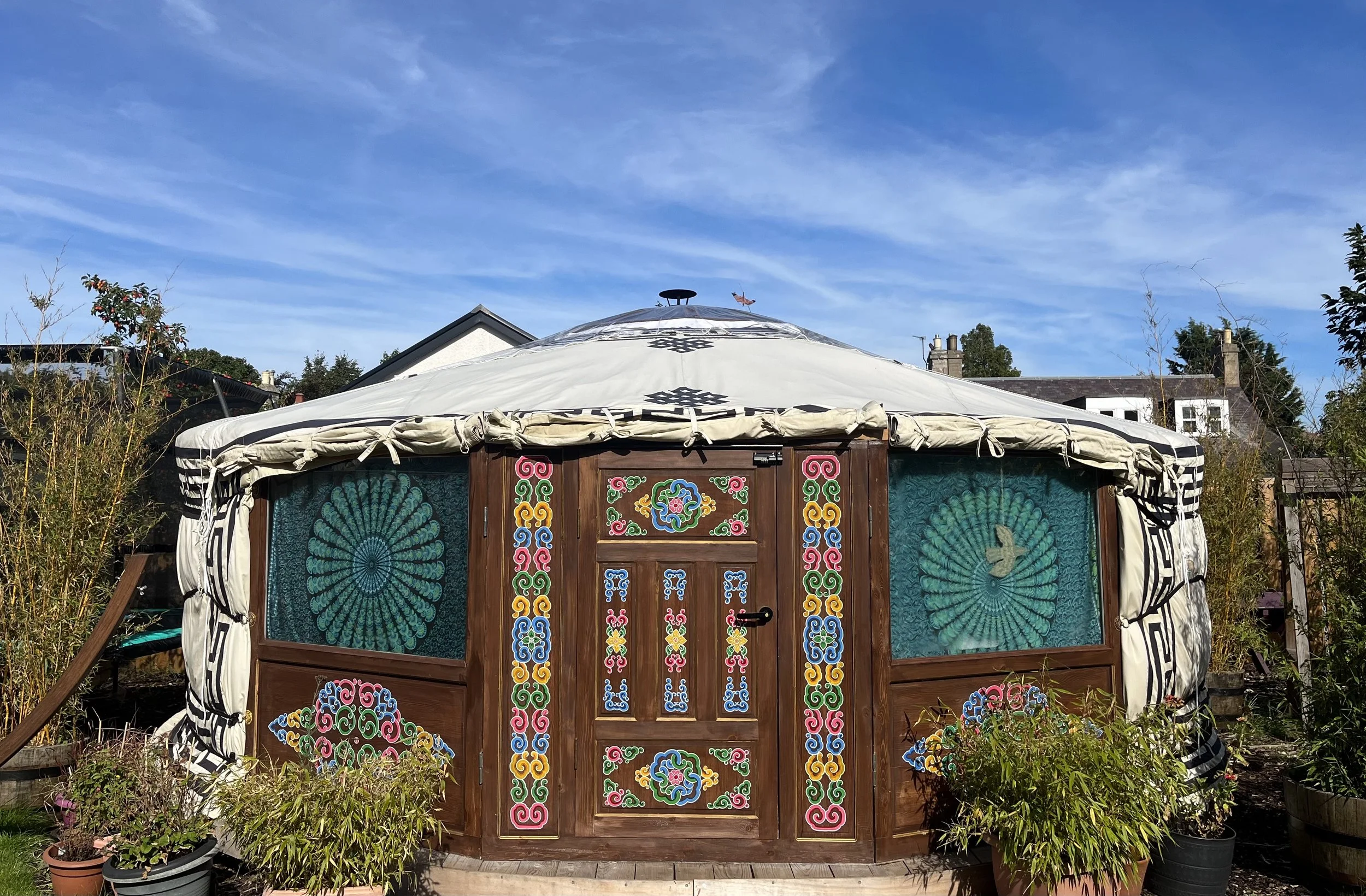 A colourful yurt with intricate paintwork on the wooden door and sides, surrounded by potted plants and located in a garden with trees under a blue sky.