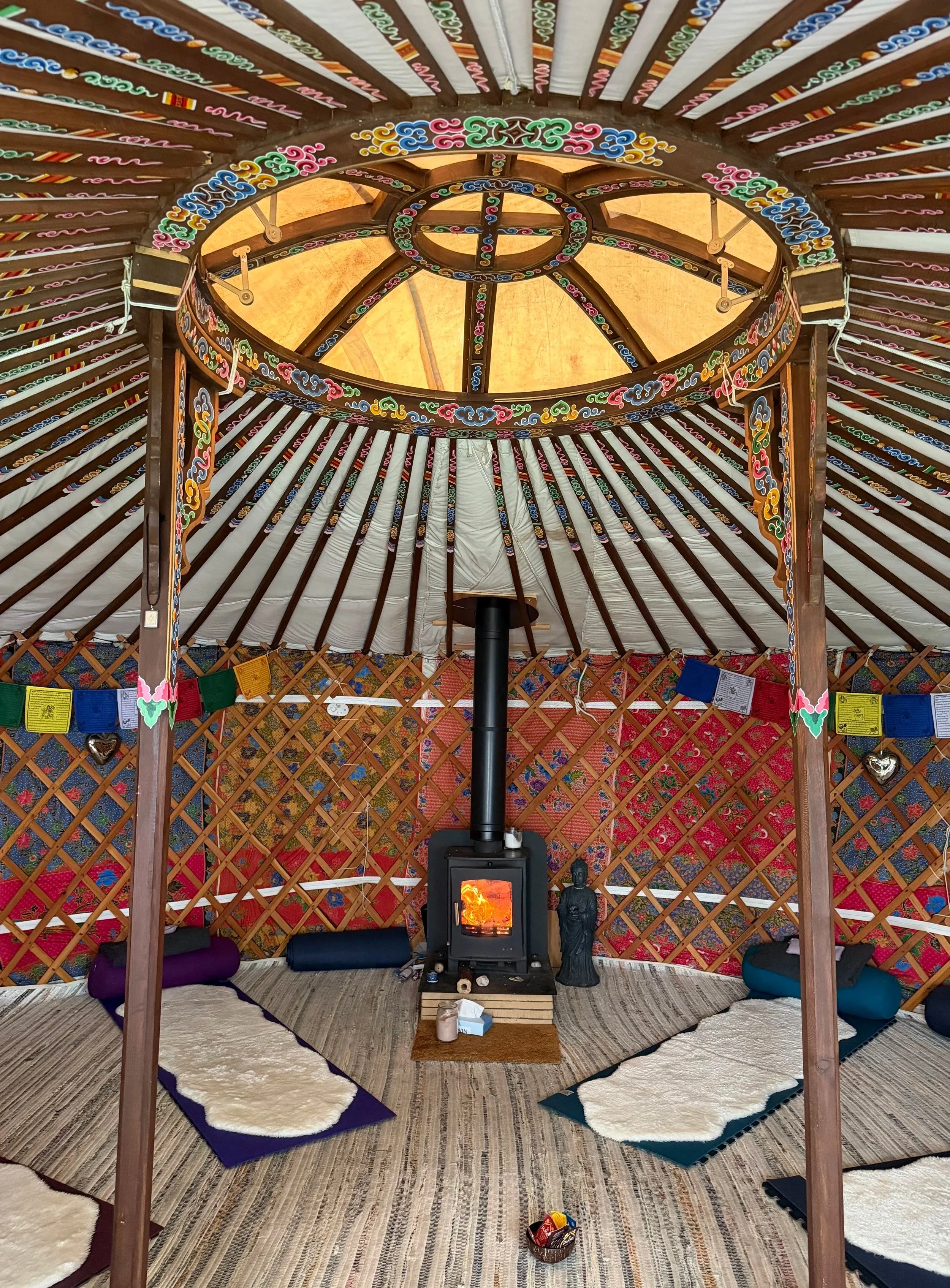 Inside a yurt with colorful, embroidered decoration, a wood stove in the center, surrounded by rugs, cushions, and prayer flags.