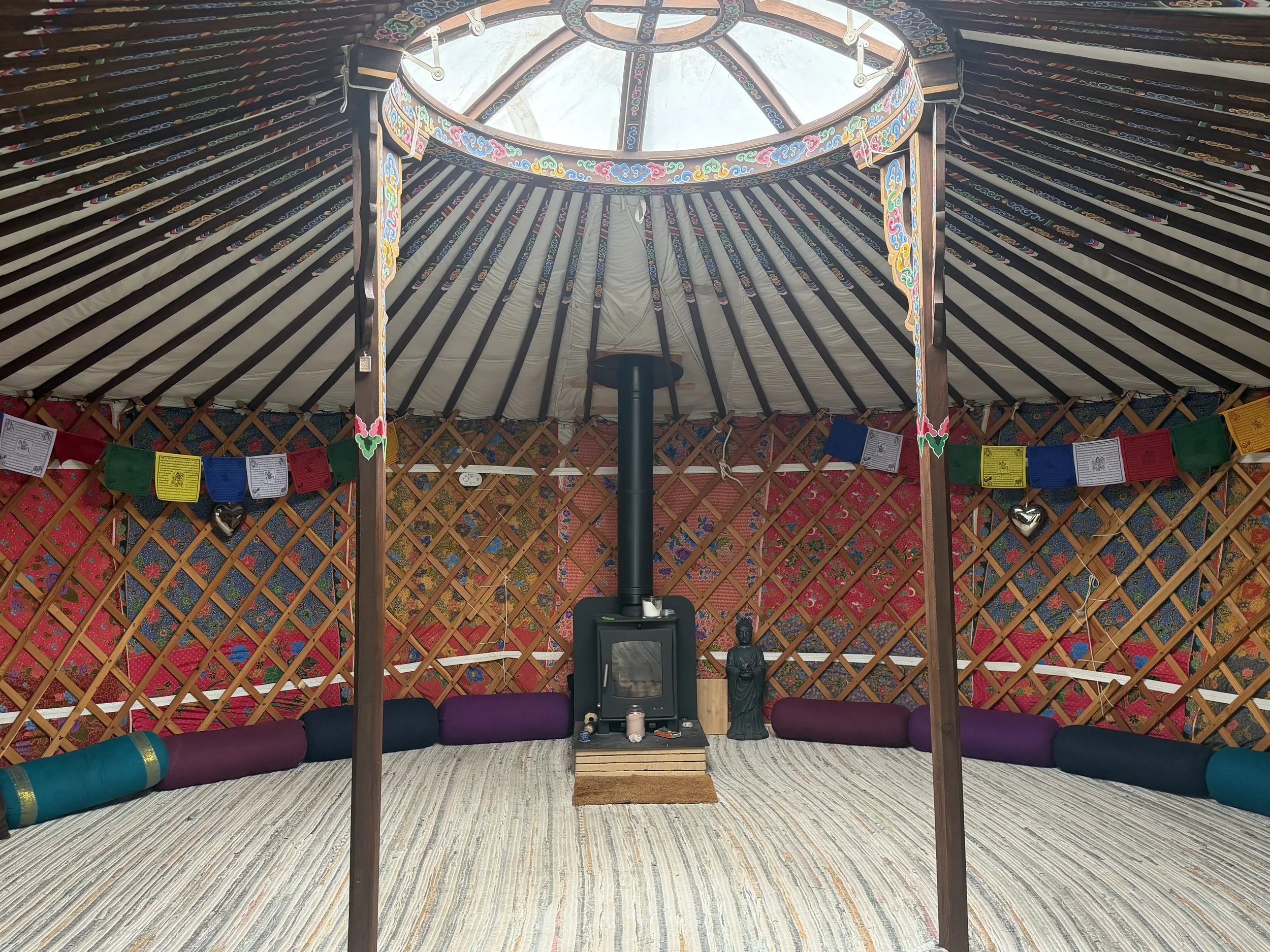 Interior of a yurt with a central wooden support structure, colorful tribal decorations and prayer flags hanging around the walls. A Buddha statue is placed on a table, with meditation cushions arranged around the space.