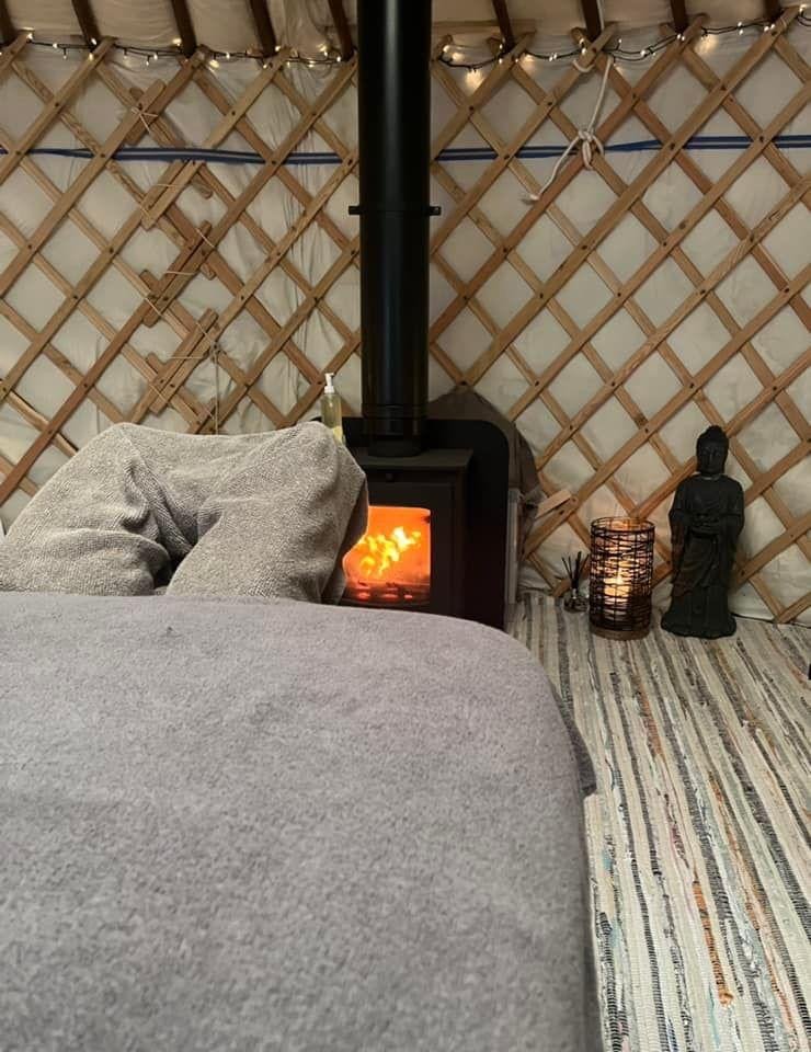 Living space with a wood stove, a gray couch, string lights, a Buddha statue, a basket with a candle, and a patterned rug on the floor. The background features a lattice wall.