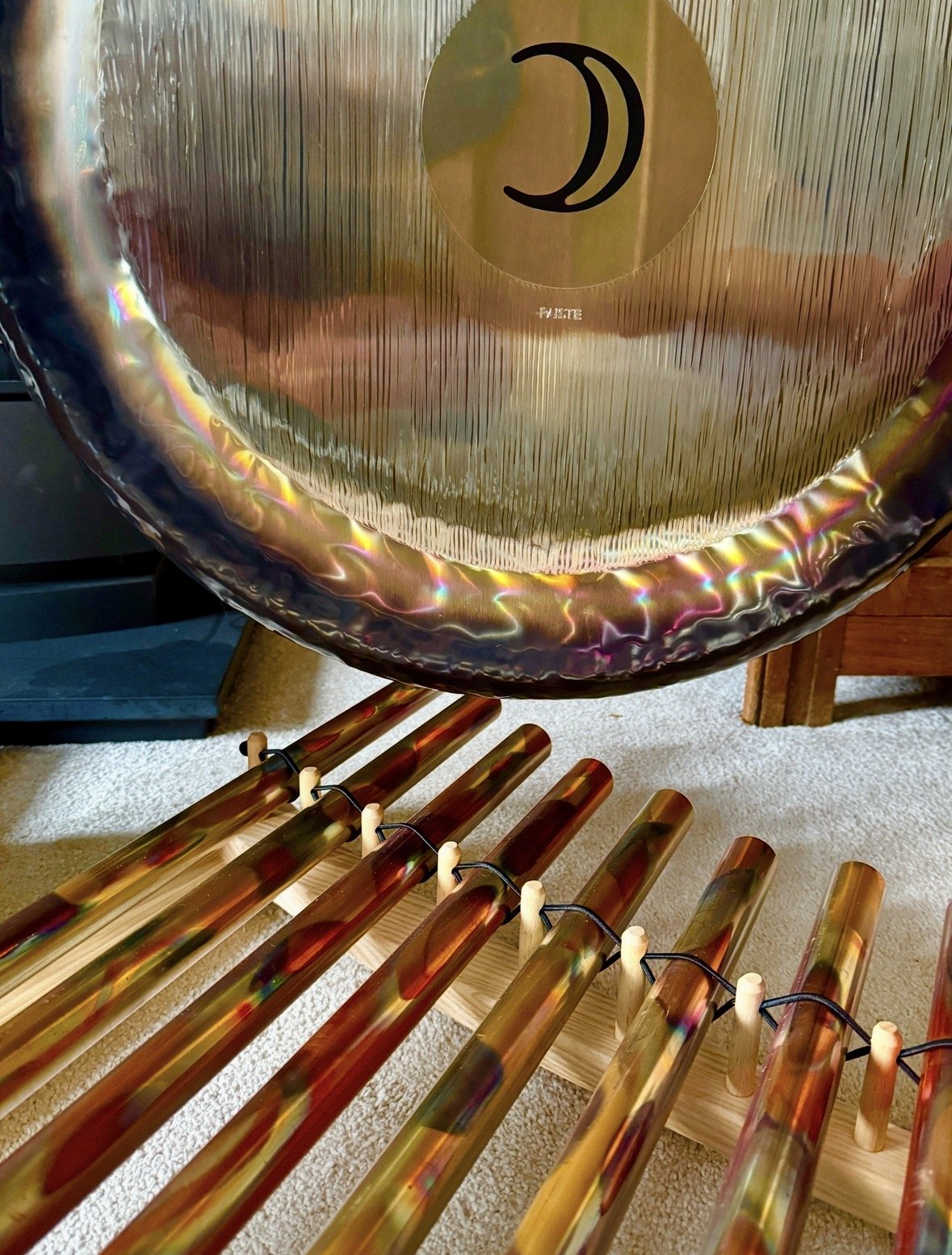 A large gong with a reflective, iridescent surface and a black sticker with a moon design. In front of it, there are several wooden sticks with a shiny, metallic finish, tied to a wooden frame, likely used as mallets to strike the gong.