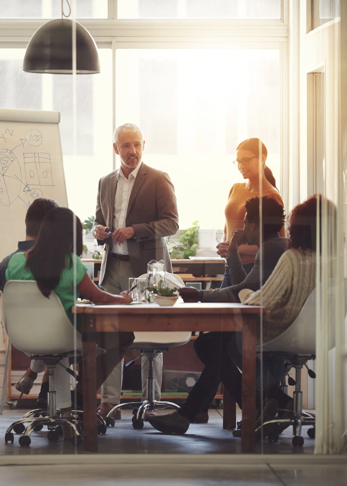 Business meeting in a modern office with diverse professionals, a man presenting in front of a whiteboard, seated colleagues at a table.