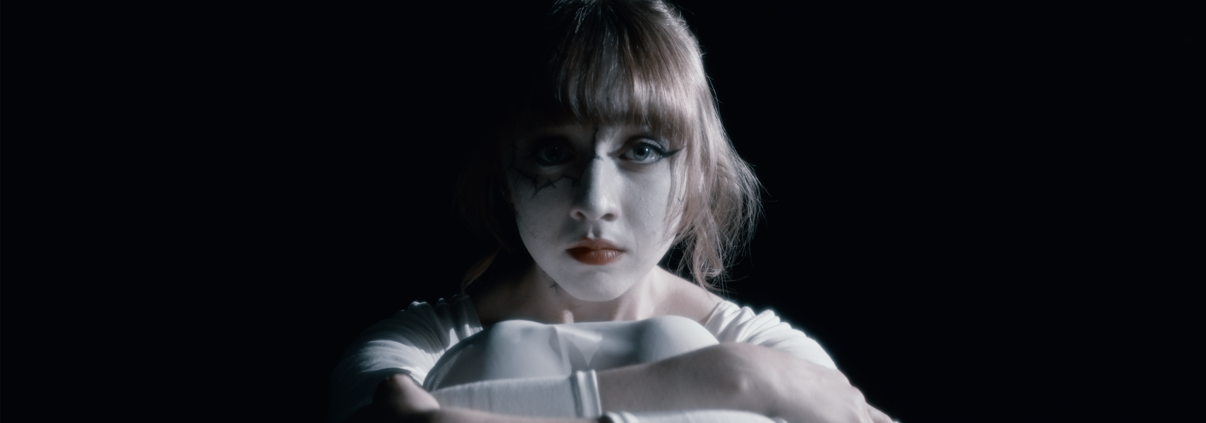 A woman with makeup resembling a clown or mime, pale face, dark eye makeup, and red lipstick, looking directly at the camera with a serious expression, arms crossed in front of her against a dark background.