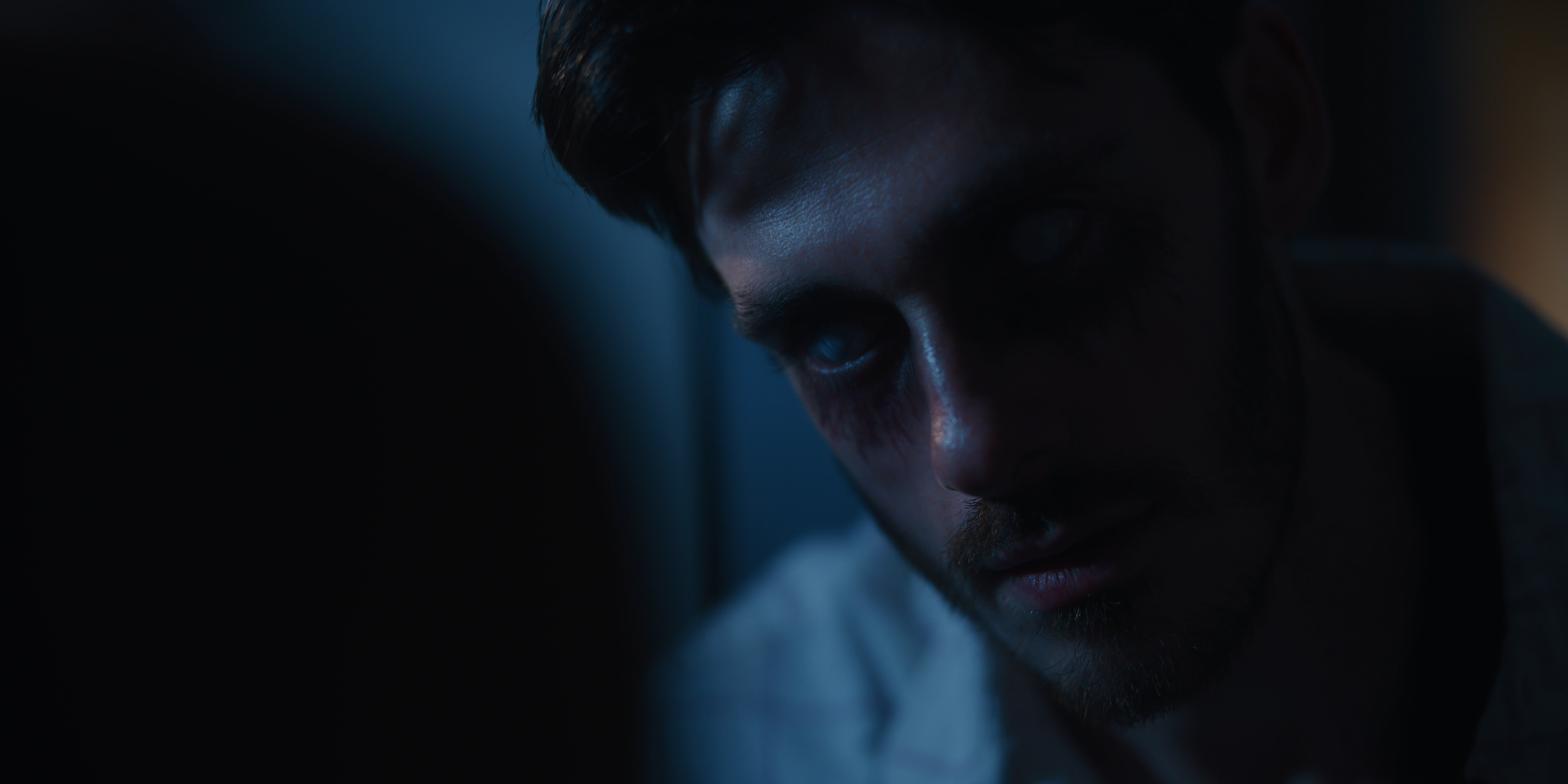 A close-up of a man with dark hair and beard, looking down in a dimly lit environment with moody lighting.