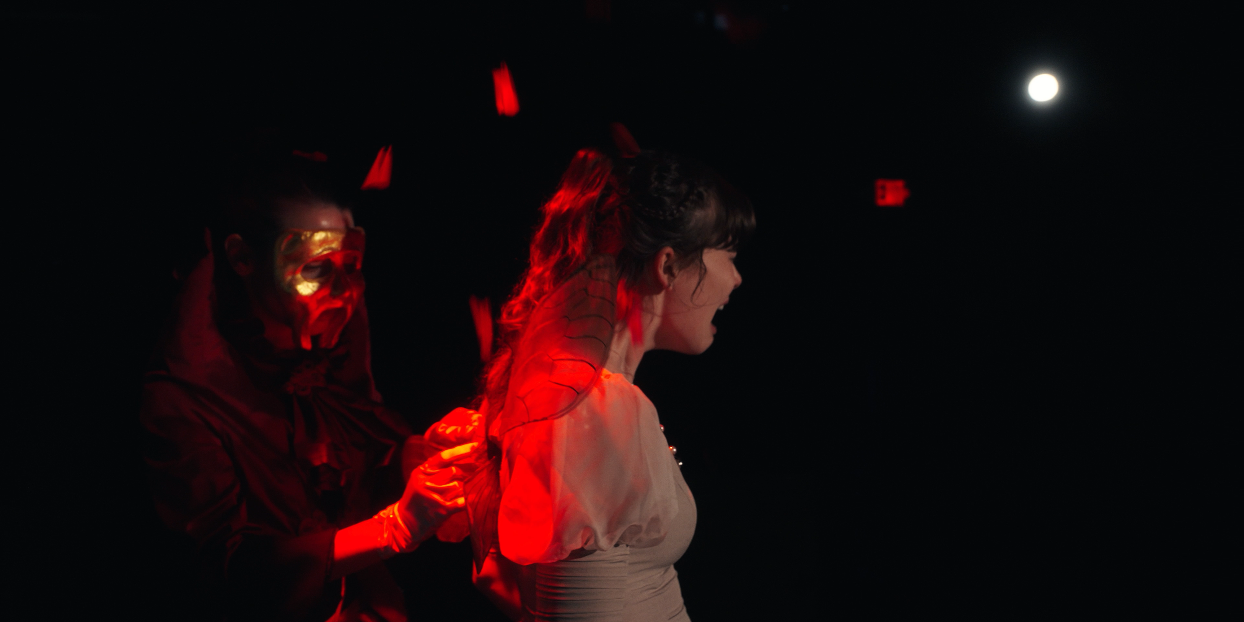 A performer dressed as a clown with a painted face, dressed in red and black, pulls on the hair of a woman with long hair who is smiling and wearing a light-colored shirt, illuminated by red stage lighting, with a moon visible in the dark night sky in the background.