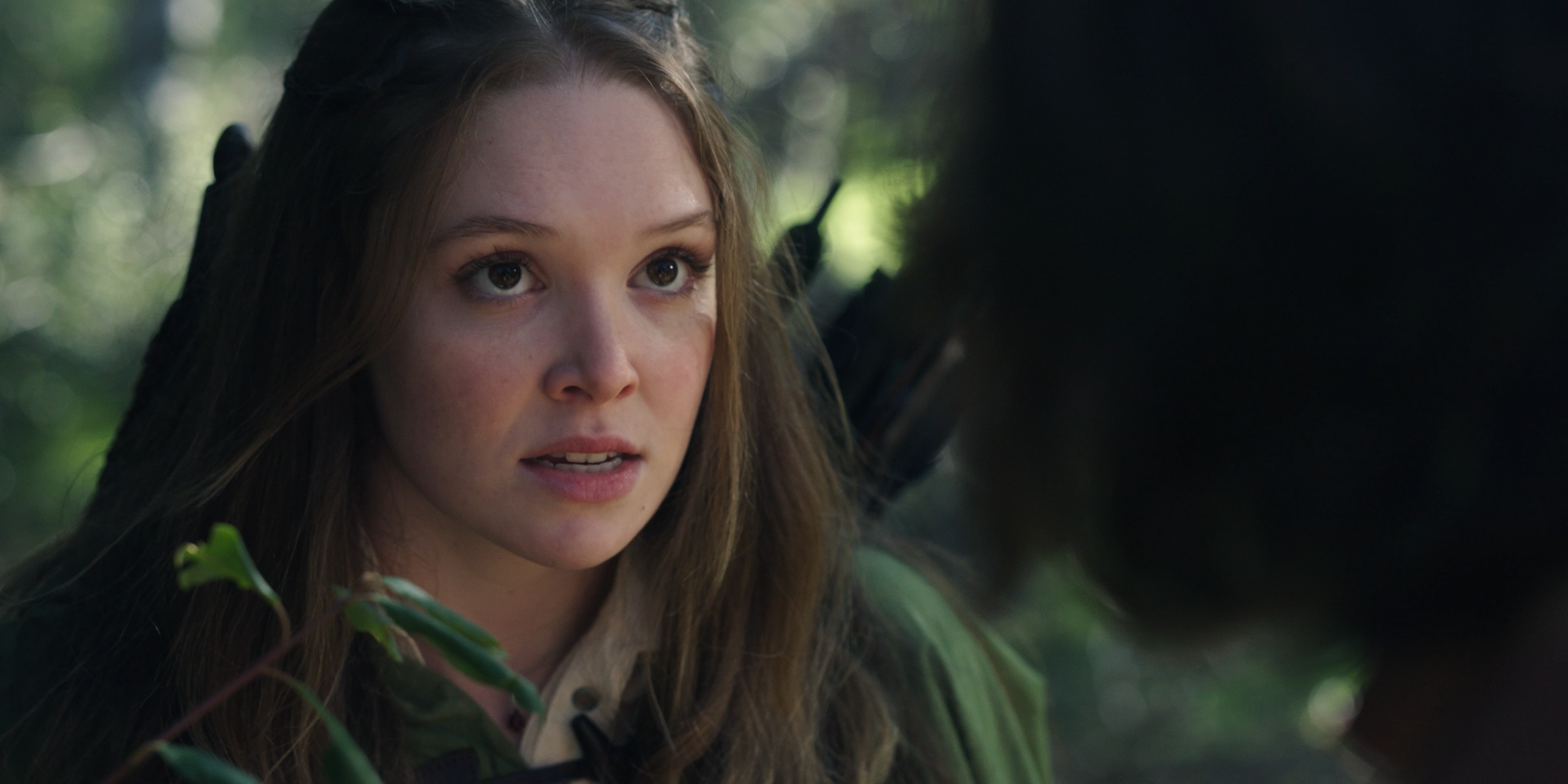 A young woman with long brown hair and brown eyes, wearing camouflage clothing, is in a forested environment, seemingly looking at something with focus.