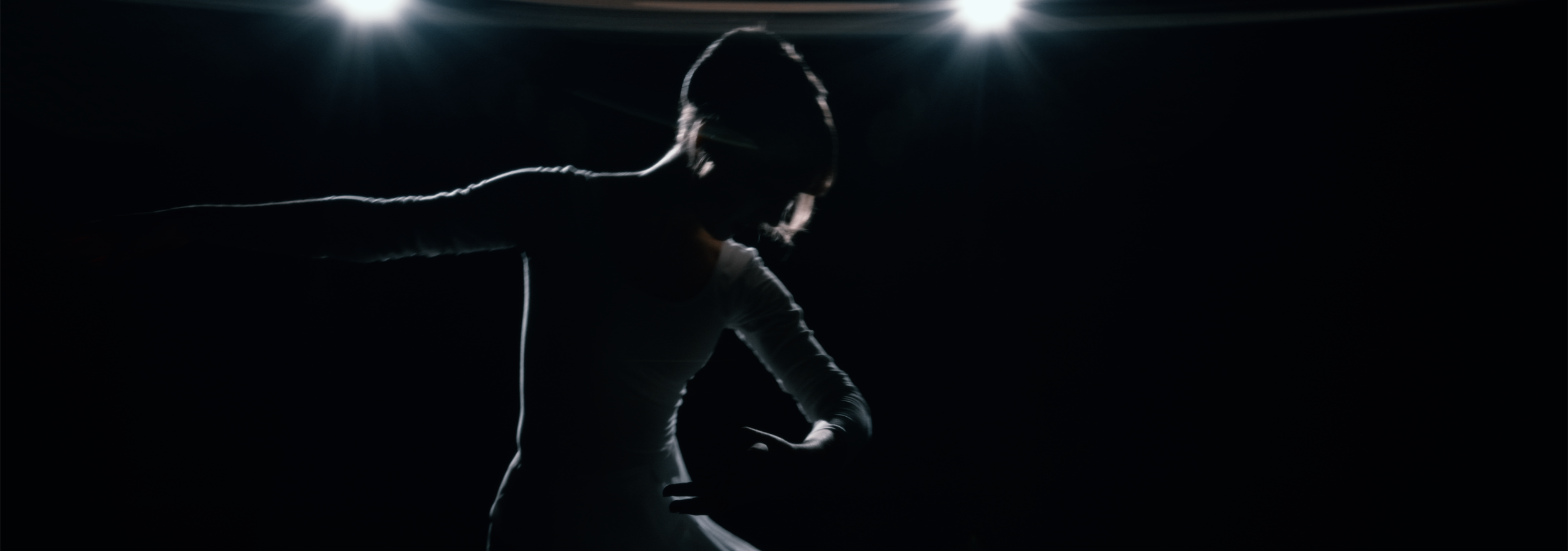 A silhouette of a woman performing a dance pose in a dark space with two bright lights behind her.