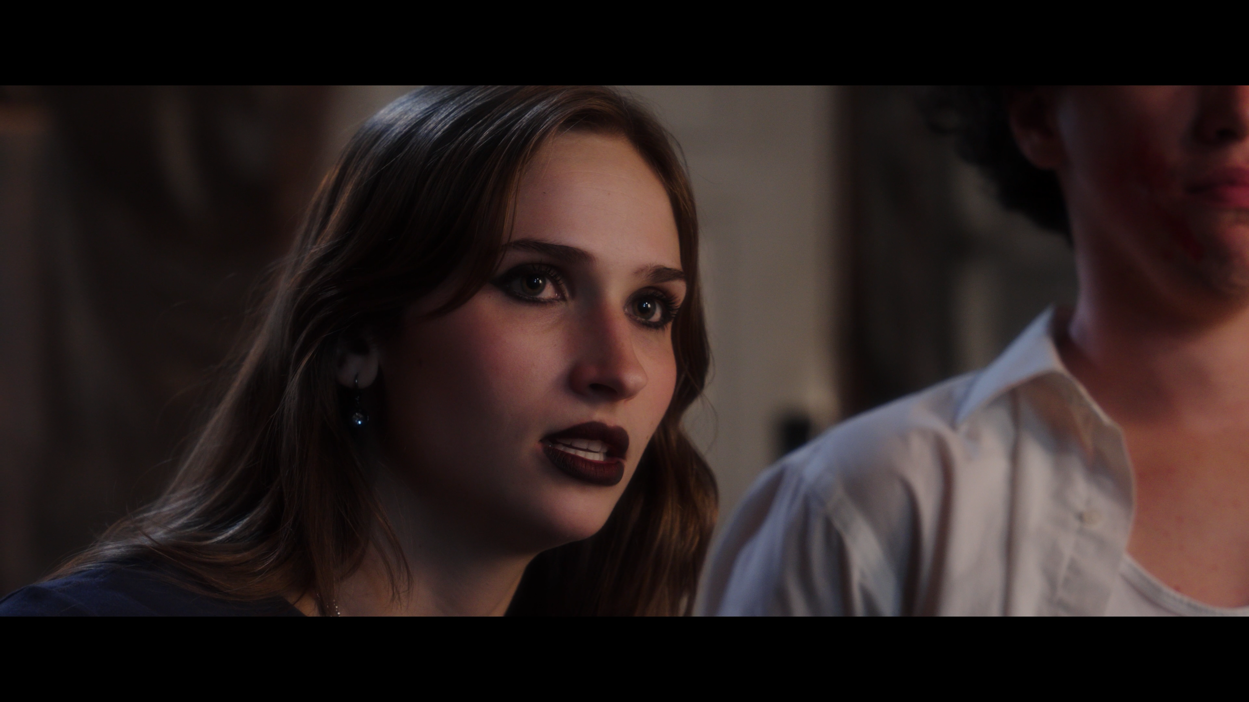A young woman with wavy brown hair, fair skin, and dark makeup appears to be speaking or reacting, with another person beside her partially visible in the image.