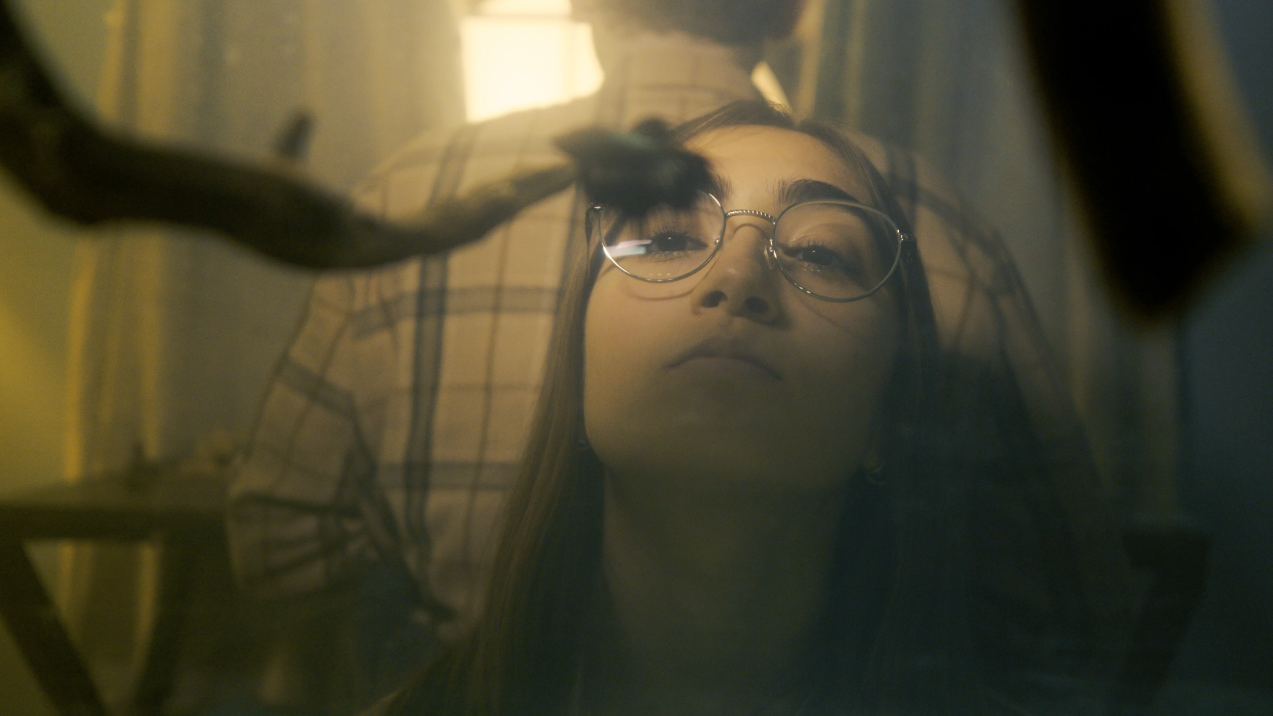 A young woman with glasses looking into a mirror, her reflection visible, with a blurred background.