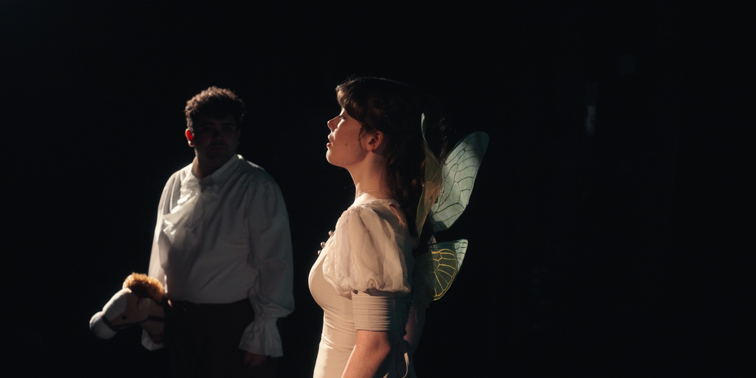 A woman with fairy wings looking to the side, and a man in a white shirt holding a stuffed animal, against a dark background.