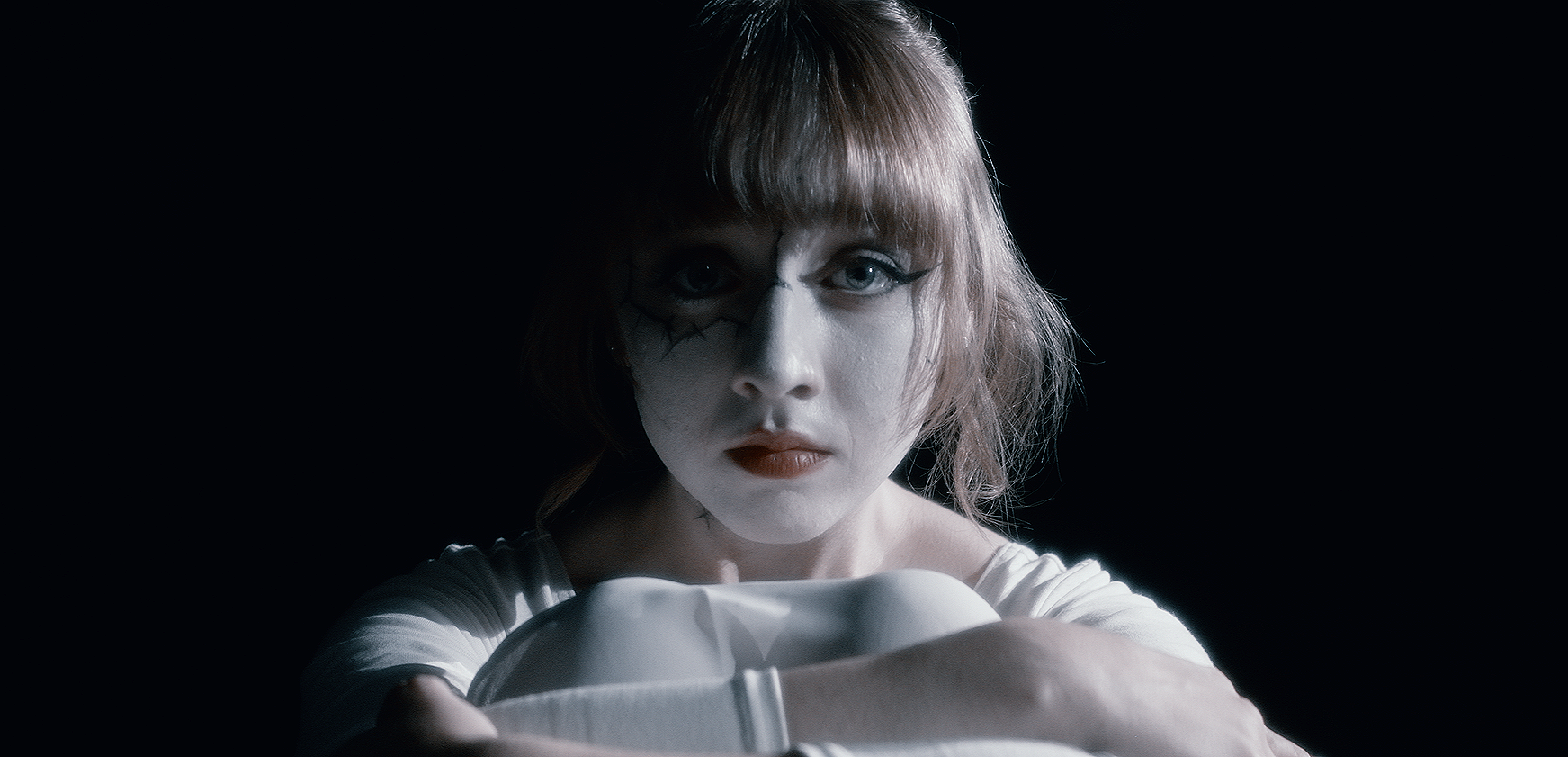 A woman with pale makeup, dark eye makeup, and red lipstick holds a white object close to her face, against a dark background.