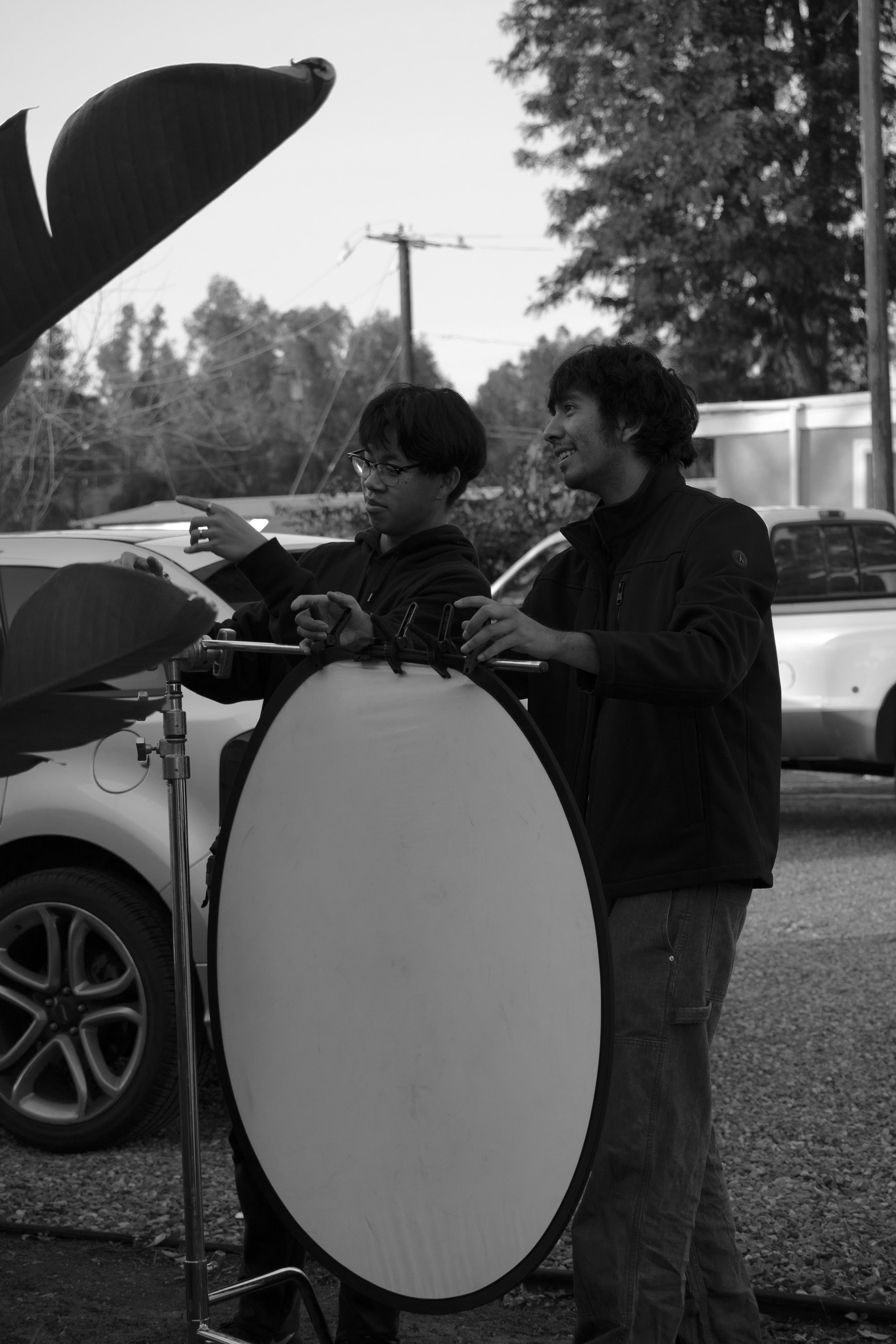 Two young men smiling and talking outdoors, one with glasses pointing at a large reflector, cars and trees in the background.