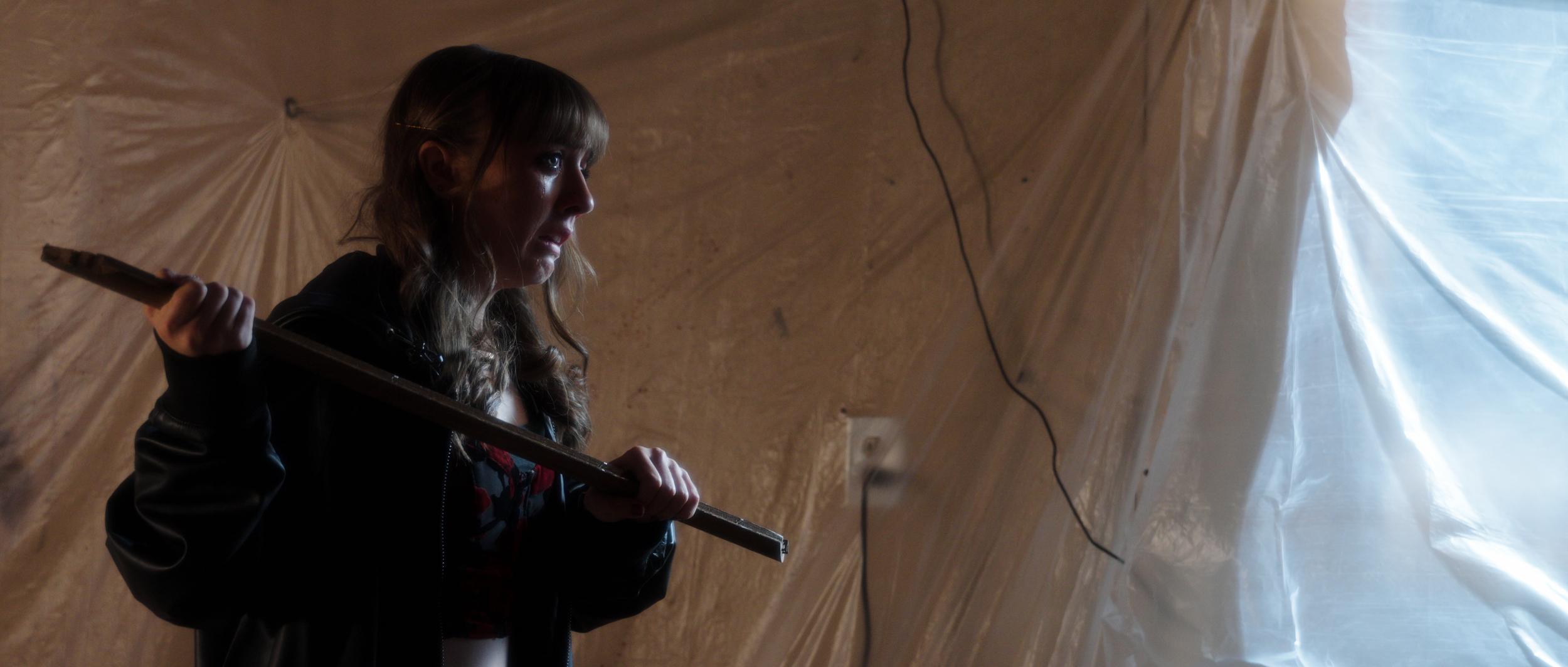A woman with long, wavy hair, wearing a black jacket, holding a wooden plank over her shoulder, standing inside a room with beige walls and a plastic sheet hanging from the ceiling.