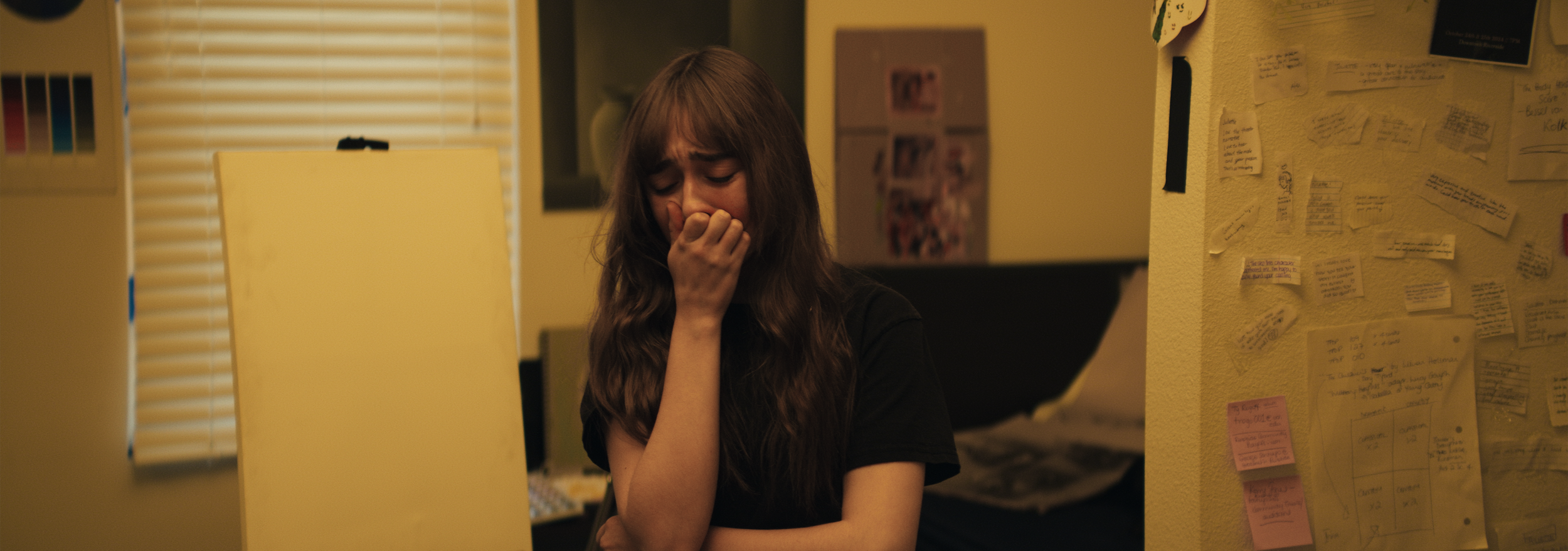 A young woman with long brown hair is standing in a room, covering her mouth with her hand, appearing upset or distressed.