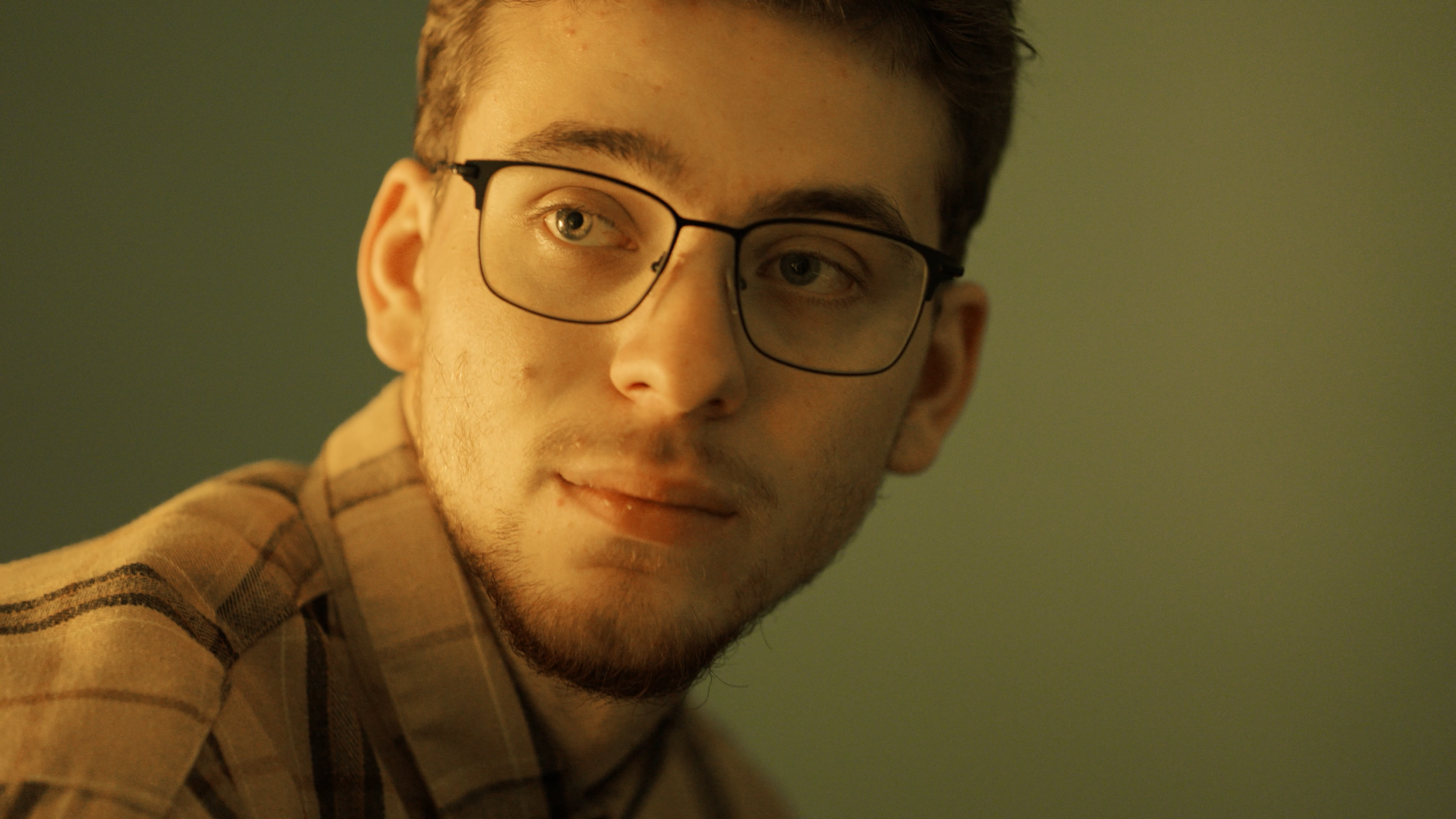 A young man with glasses and facial hair looking to the side, wearing a plaid shirt.