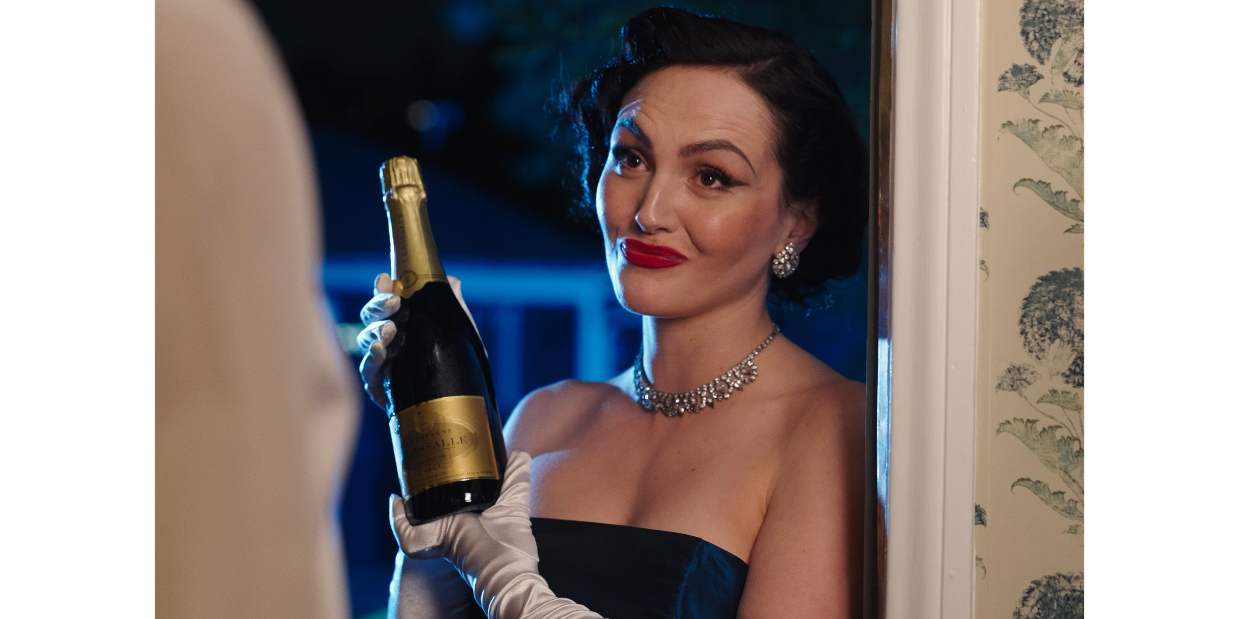 A woman with vintage styling, wearing a black strapless dress, diamond earrings, and a diamond necklace, is smiling and holding a bottle of champagne while looking at someone.