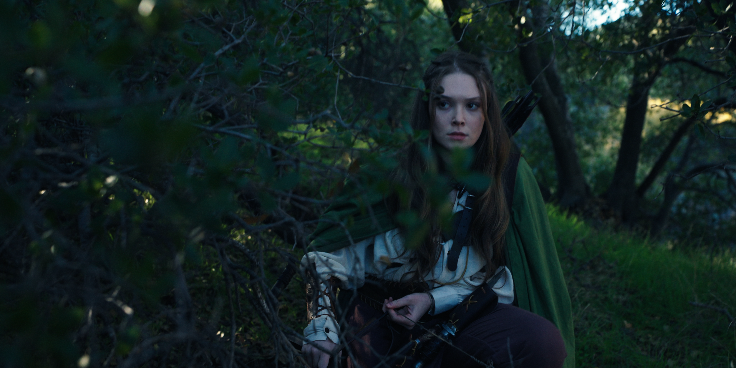 A young woman with long brown hair, dressed in outdoor gear, sits in a forest among dense green foliage, holding a bow and quiver of arrows, with a serious expression.
