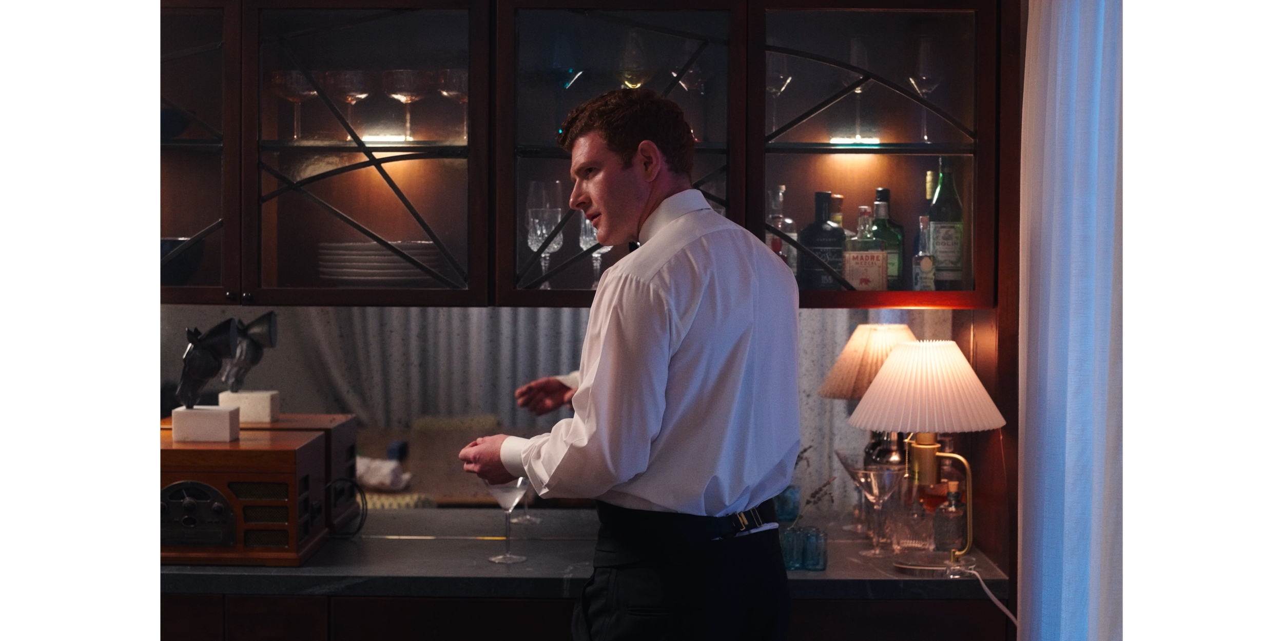 A man in a white dress shirt and black pants standing in a dimly lit room, preparing a drink at a bar with liquor bottles and glassware in the background, and a small table lamp on the side.