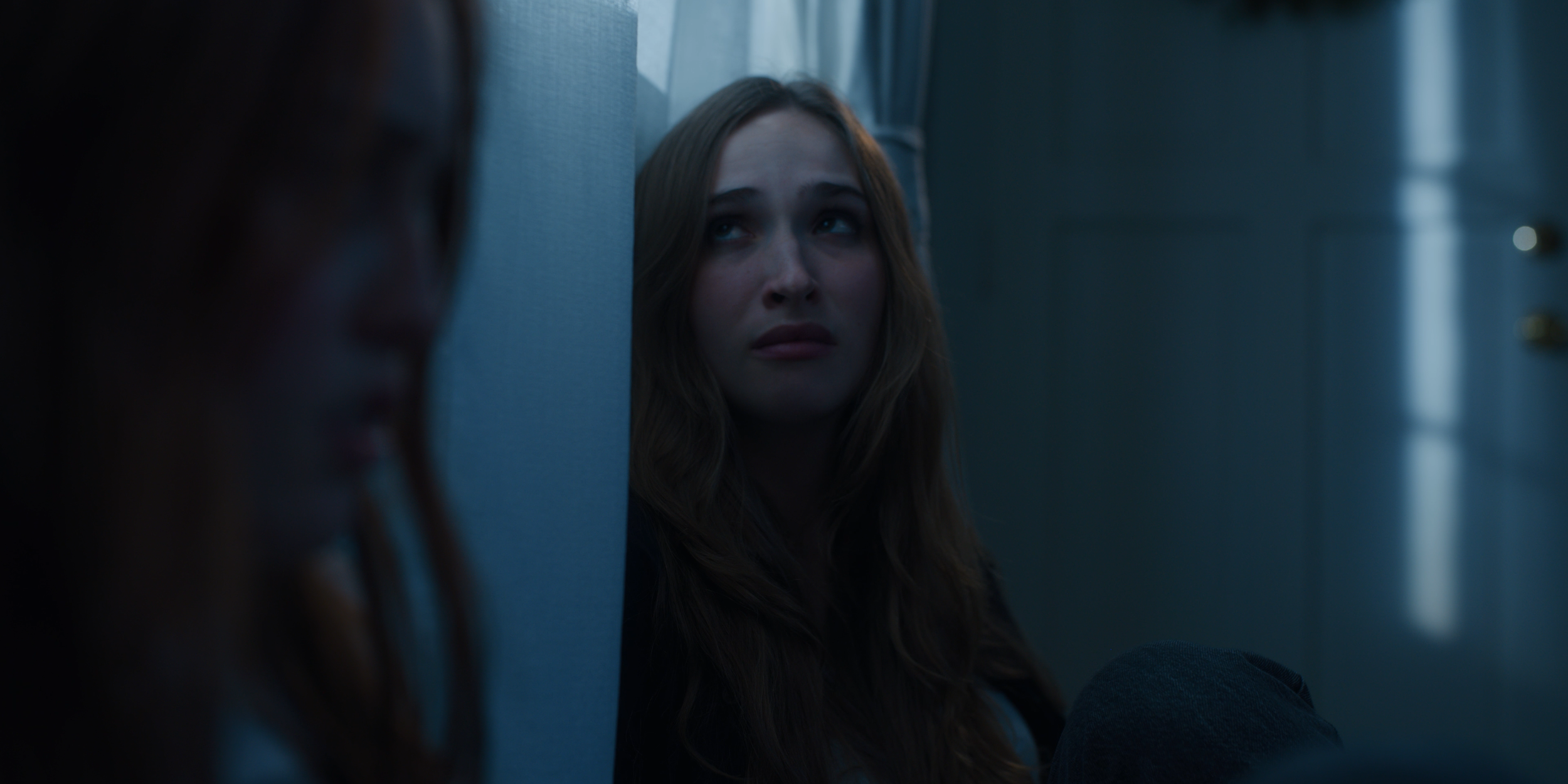 A woman with long brown hair sitting against a wall, looking distressed or sad, with dim lighting creating a somber mood.