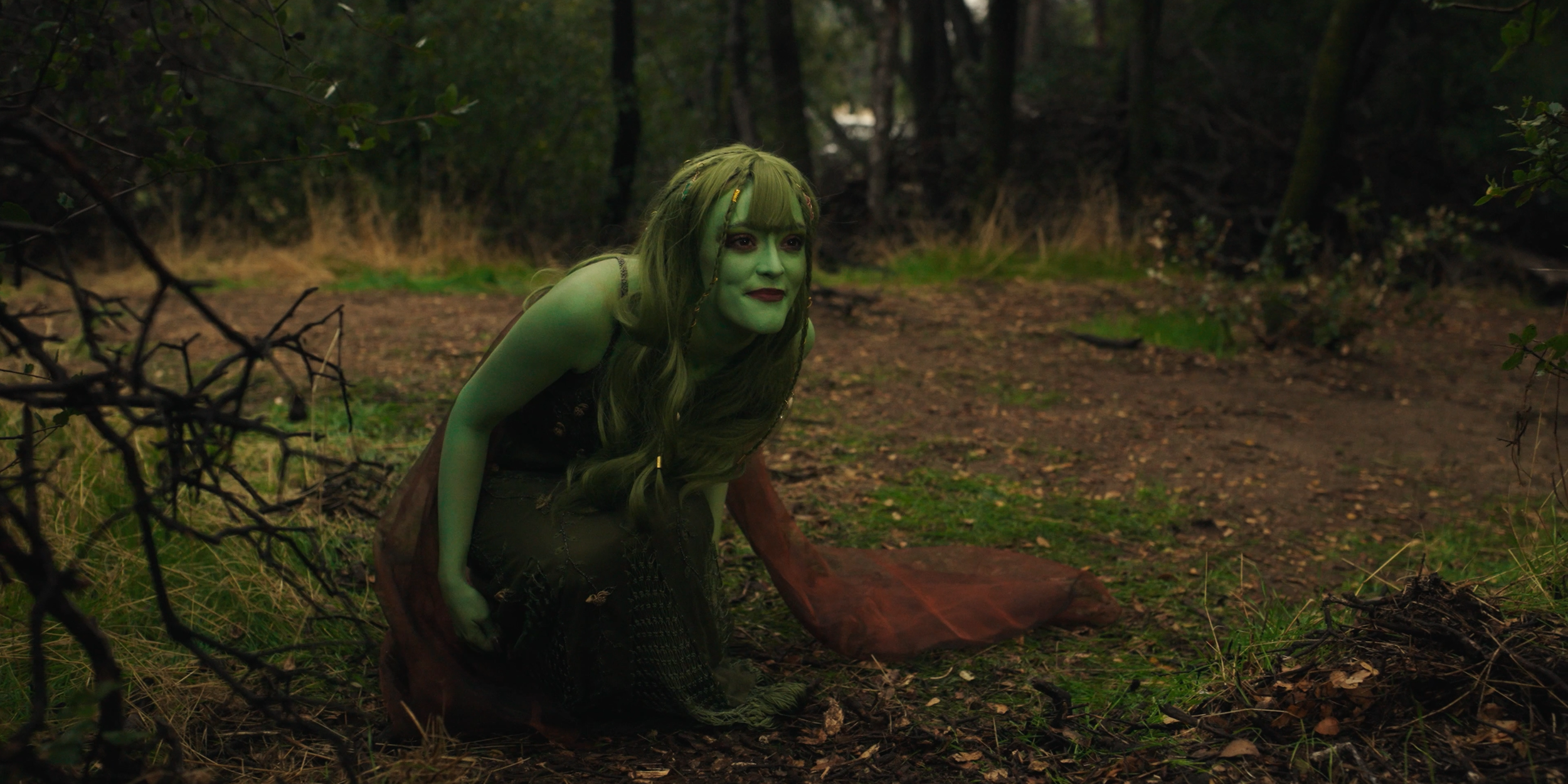 A person with green face and green hair dressed in a fantasy costume, crouching with a curious expression in a forest setting.