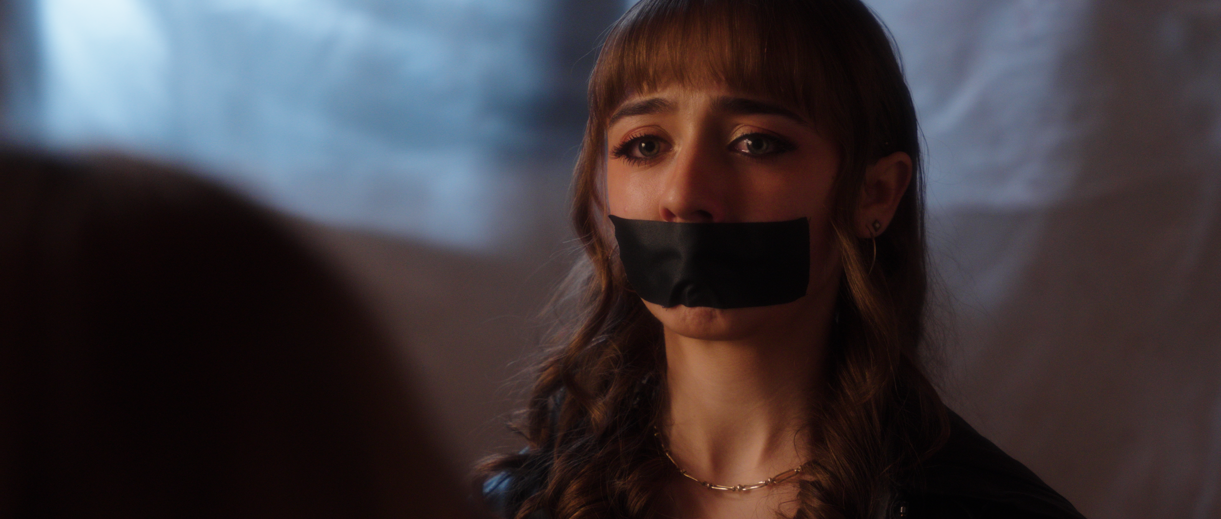 A young woman with brown hair and light-colored eyes is shown with black tape over her mouth, looking serious, in a dimly lit environment with a dark gray background.