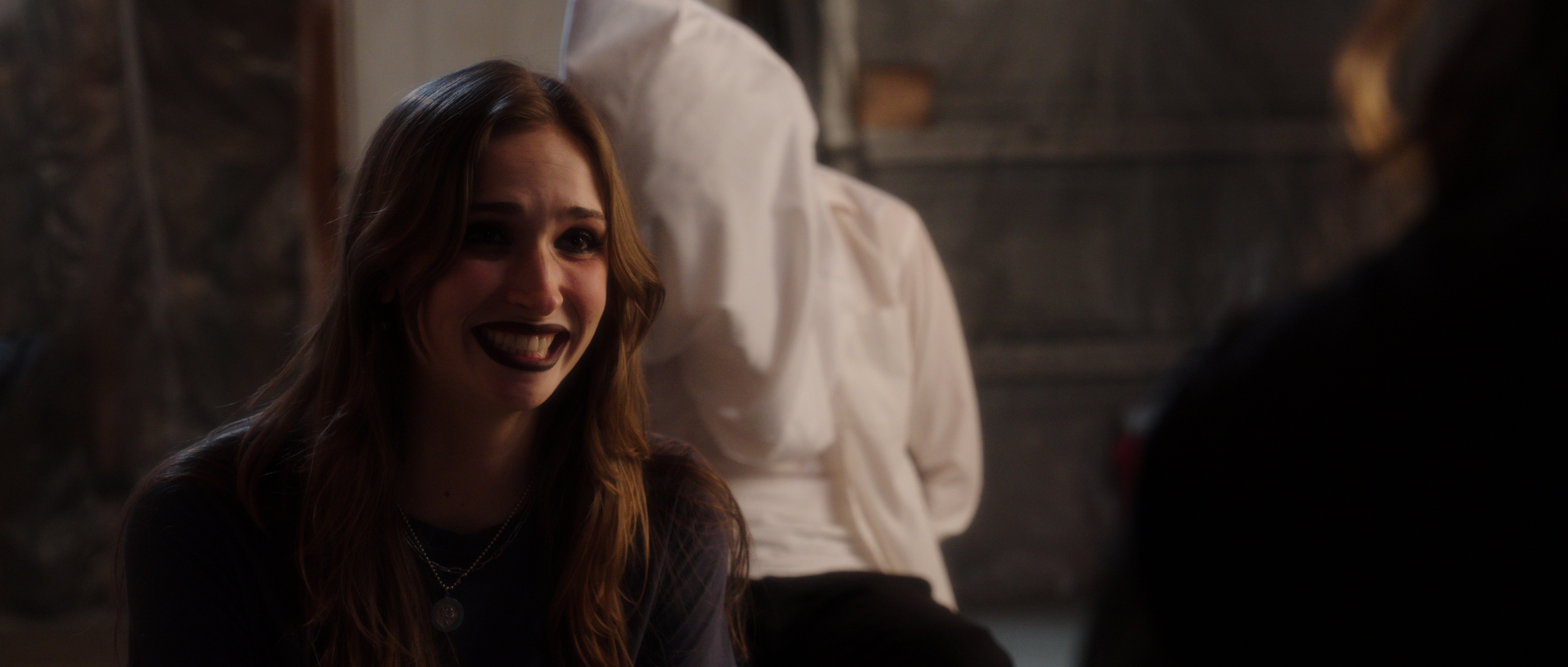 A young woman with long red hair smiling and talking, seated indoors with a stone wall in the background.