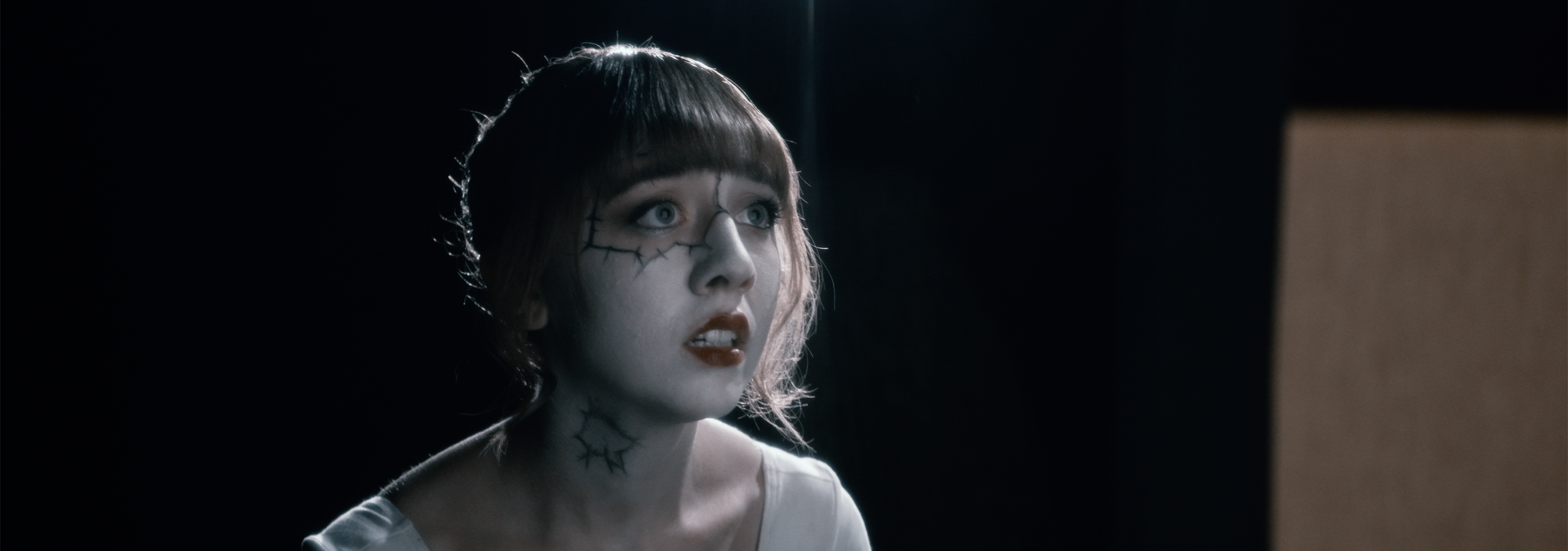 A woman with pale skin and short brown hair with bangs, makeup with stitches drawn on her face, looking at a computer screen with a surprised or shocked expression, in a dark room