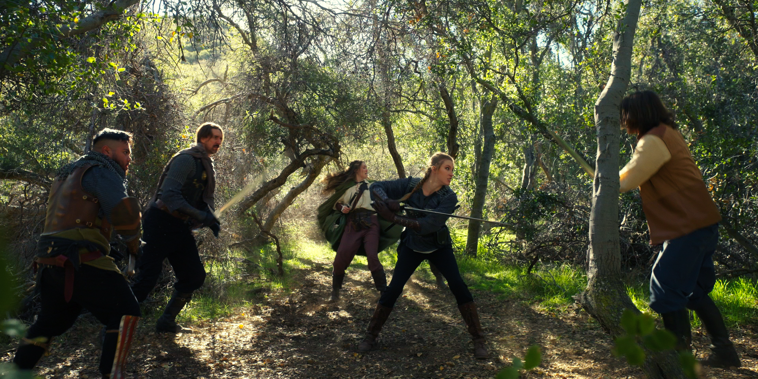 Group of five people engaged in a sword fight in a dense forest during daytime.