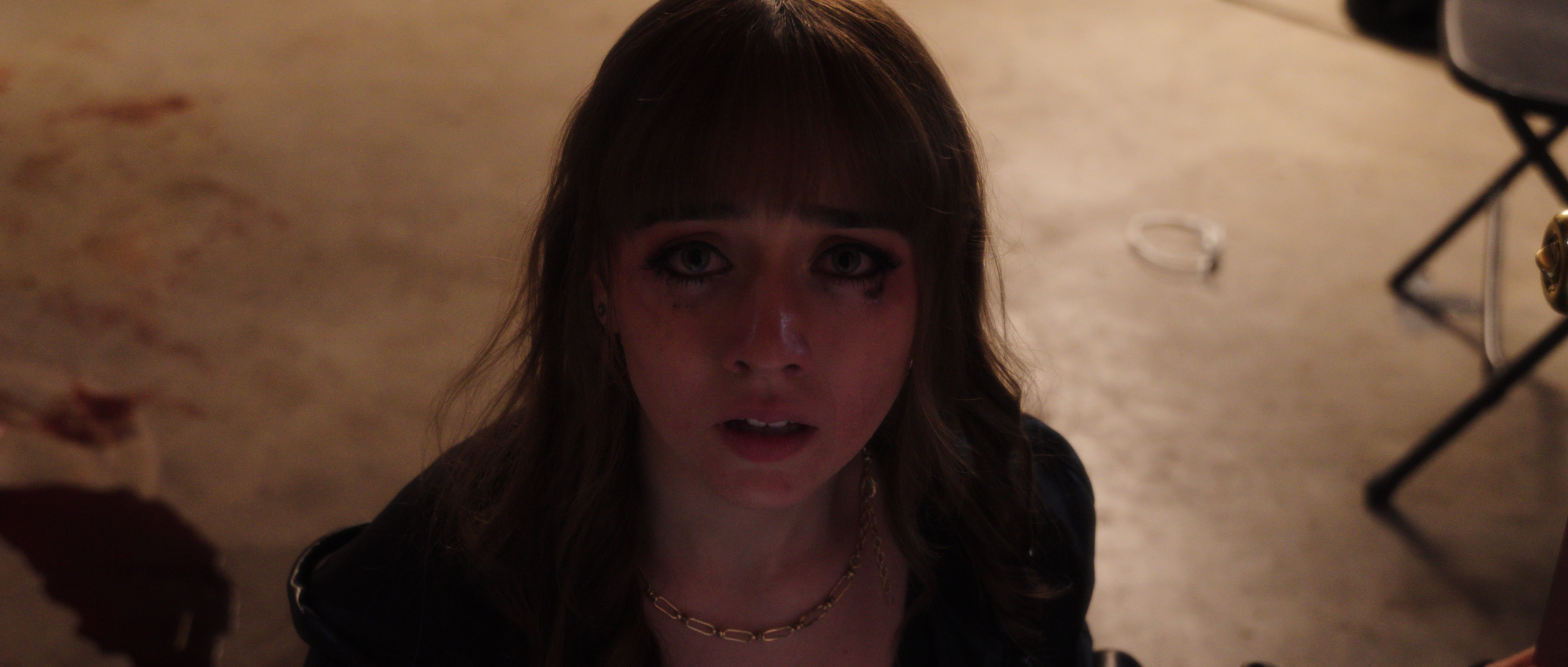 A young woman with long brown hair and blue eyes, wearing dark makeup and jewelry, looking up at the camera with a surprised or distressed expression.