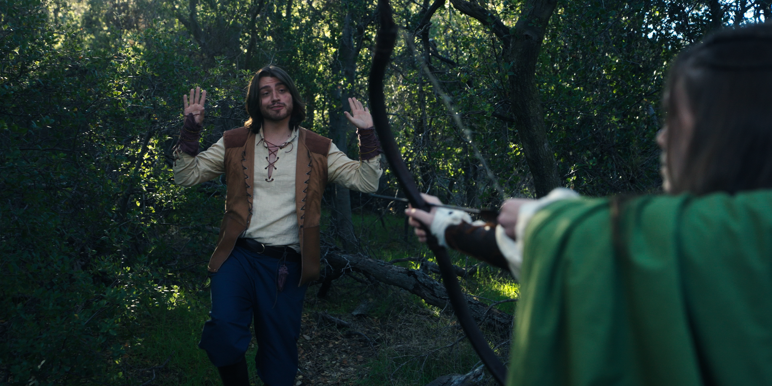 Two individuals in a forest; one person with long hair and medieval-style clothing holds their hands up as if surrendering while the other person, partially visible, points a bow at them.