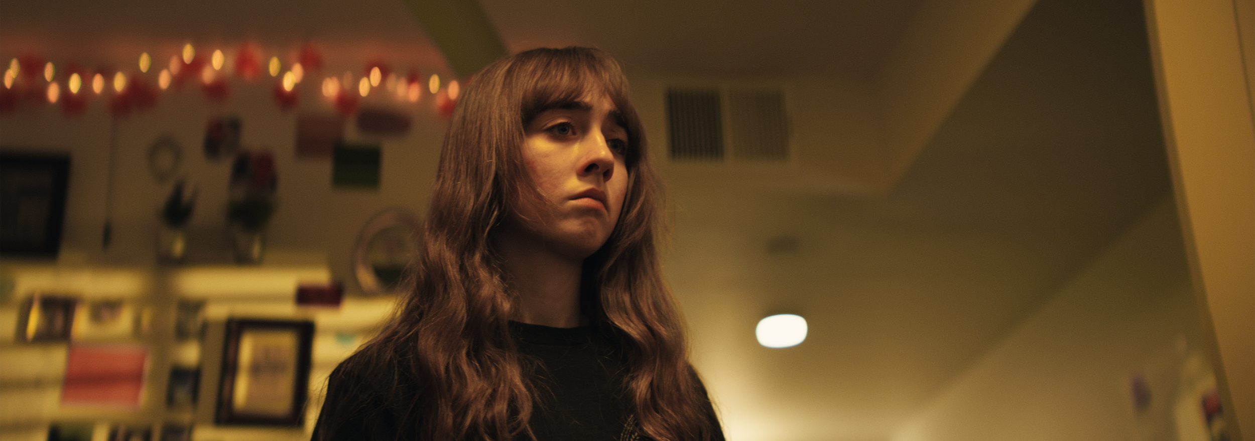 Young woman with long wavy brown hair looking to the side in a dimly lit room with blurry decorations on the wall and warm lighting.